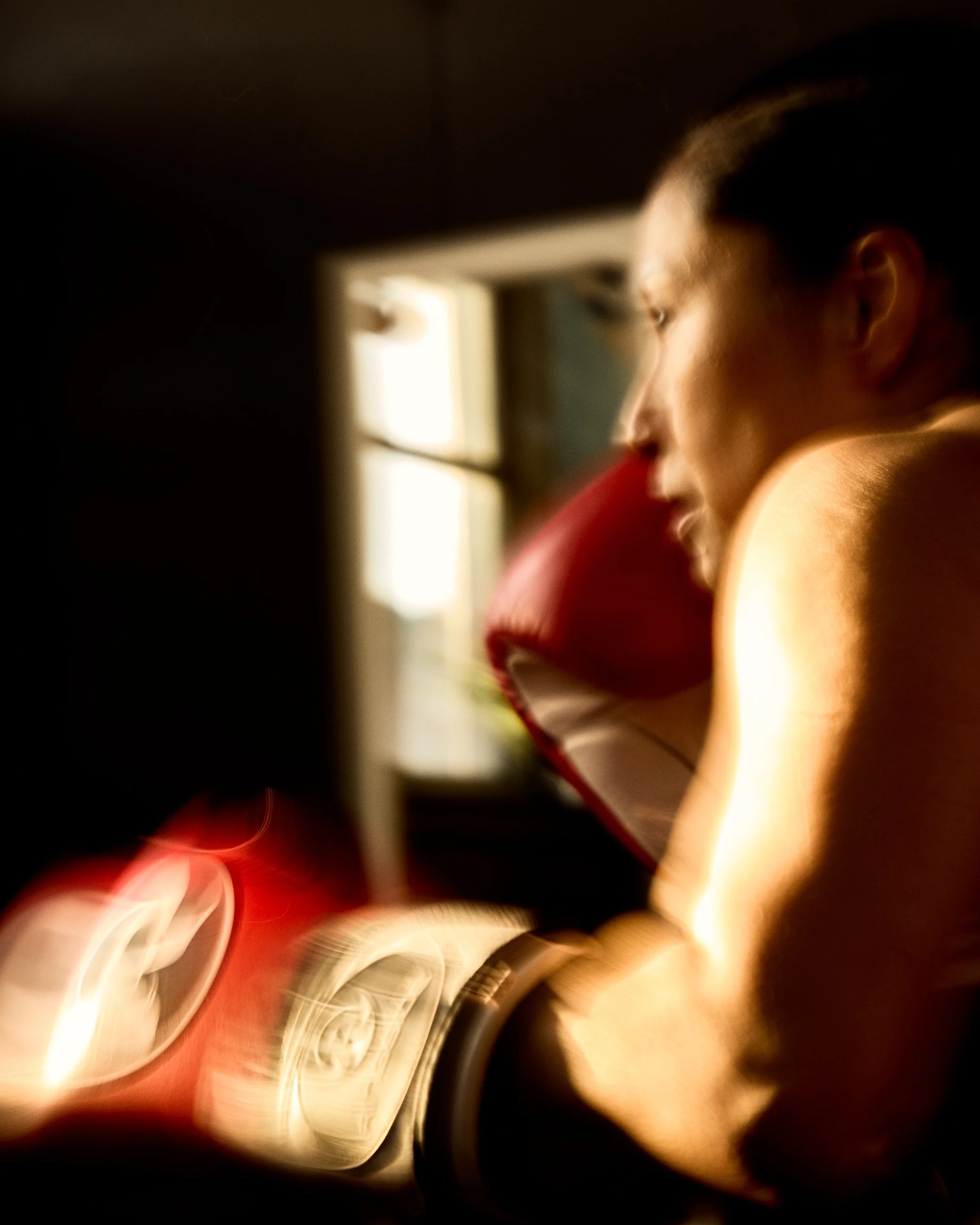 A woman in profile, wearing boxing gloves, practicing boxing in a dimly lit room with sunlight coming through a window.