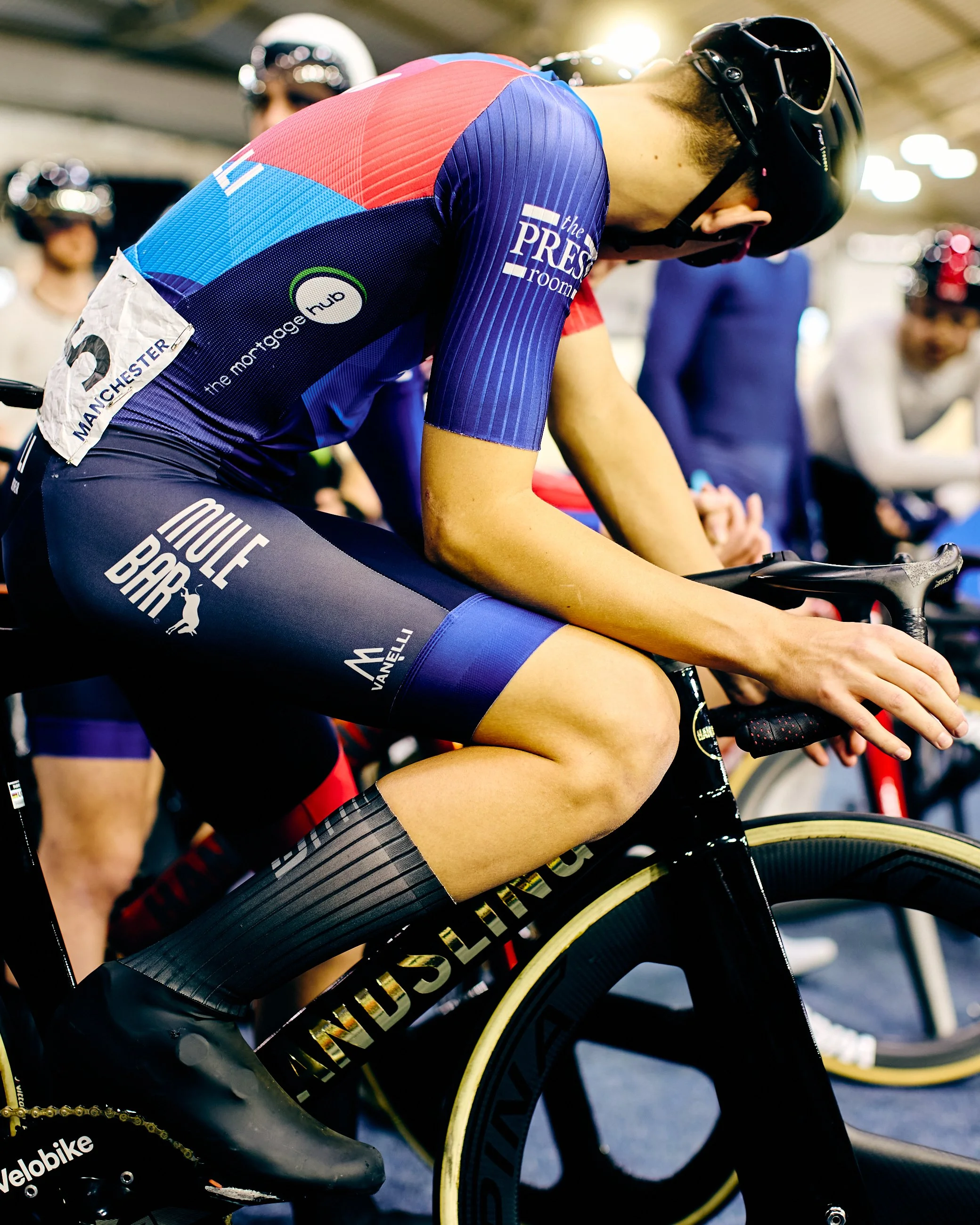 A cyclist in a racing kit with sponsors' logos, wearing a helmet and sunglasses, rides a black aerodynamic bicycle indoors, with other cyclists and spectators in the background.