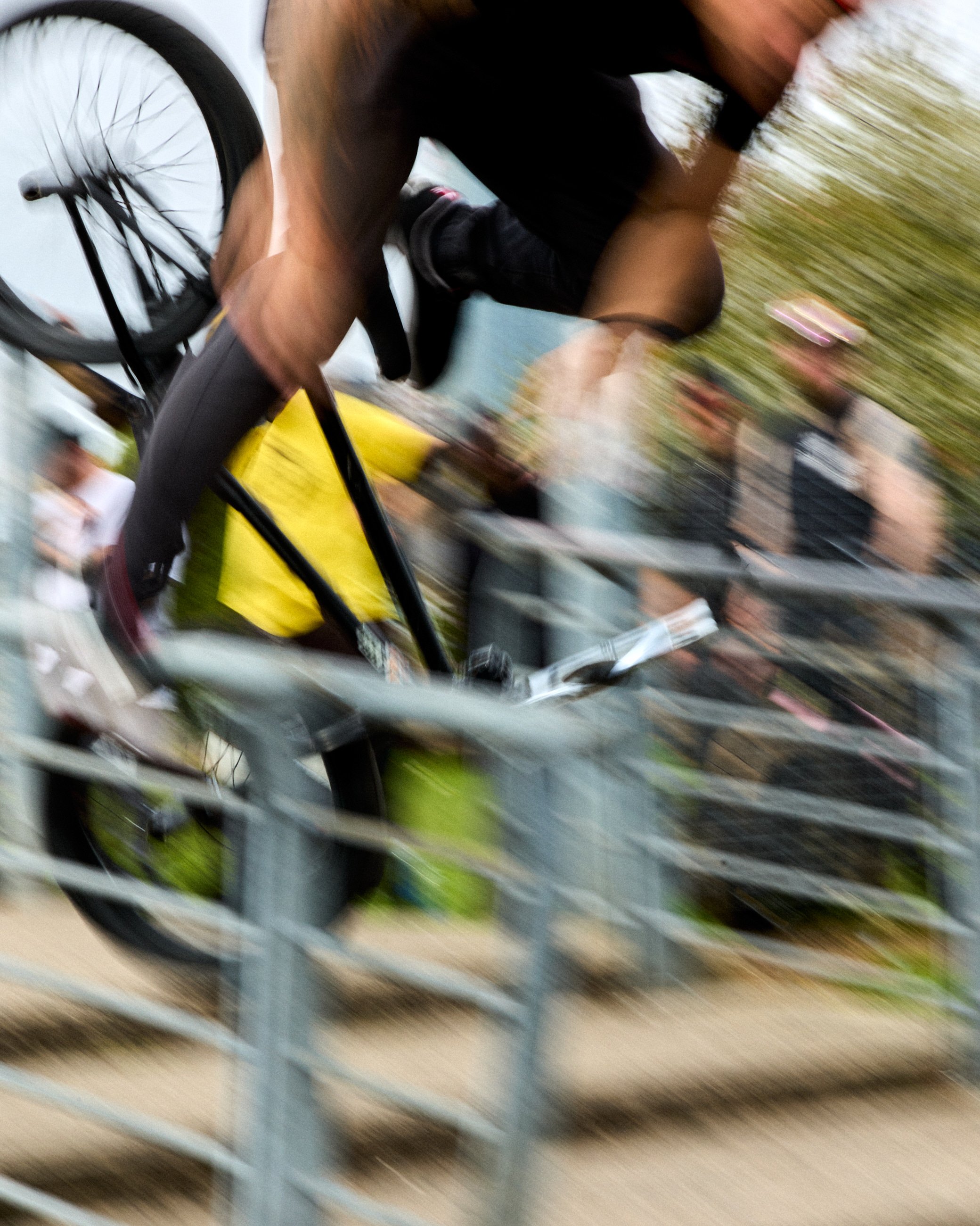 A person on a bicycle is captured in motion, leaning forward as they move past a metal fence. The background shows blurred outdoor scenery with trees and buildings.