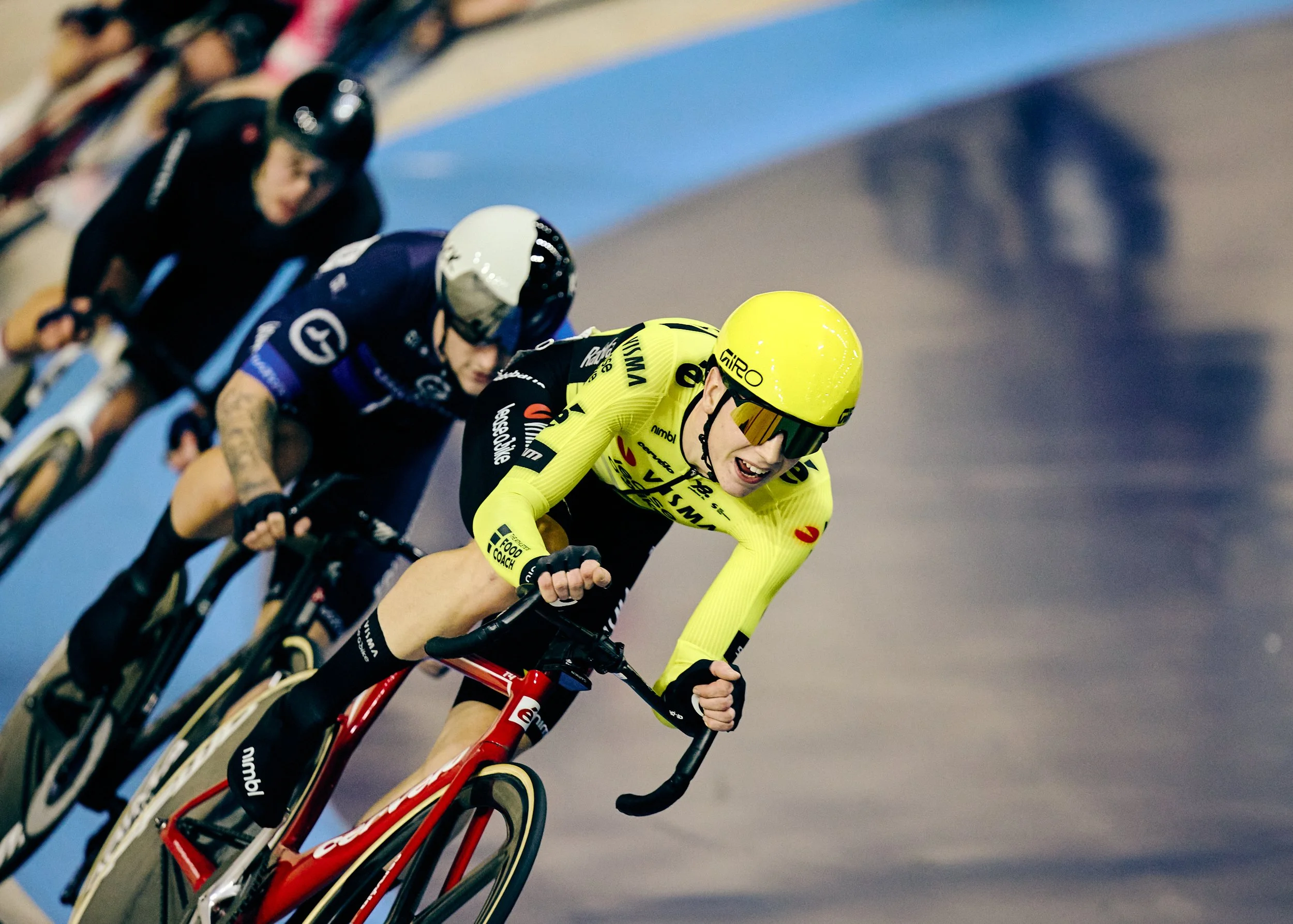 Cyclists racing on an indoor velodrome track, with one cyclist in yellow leading and others behind.