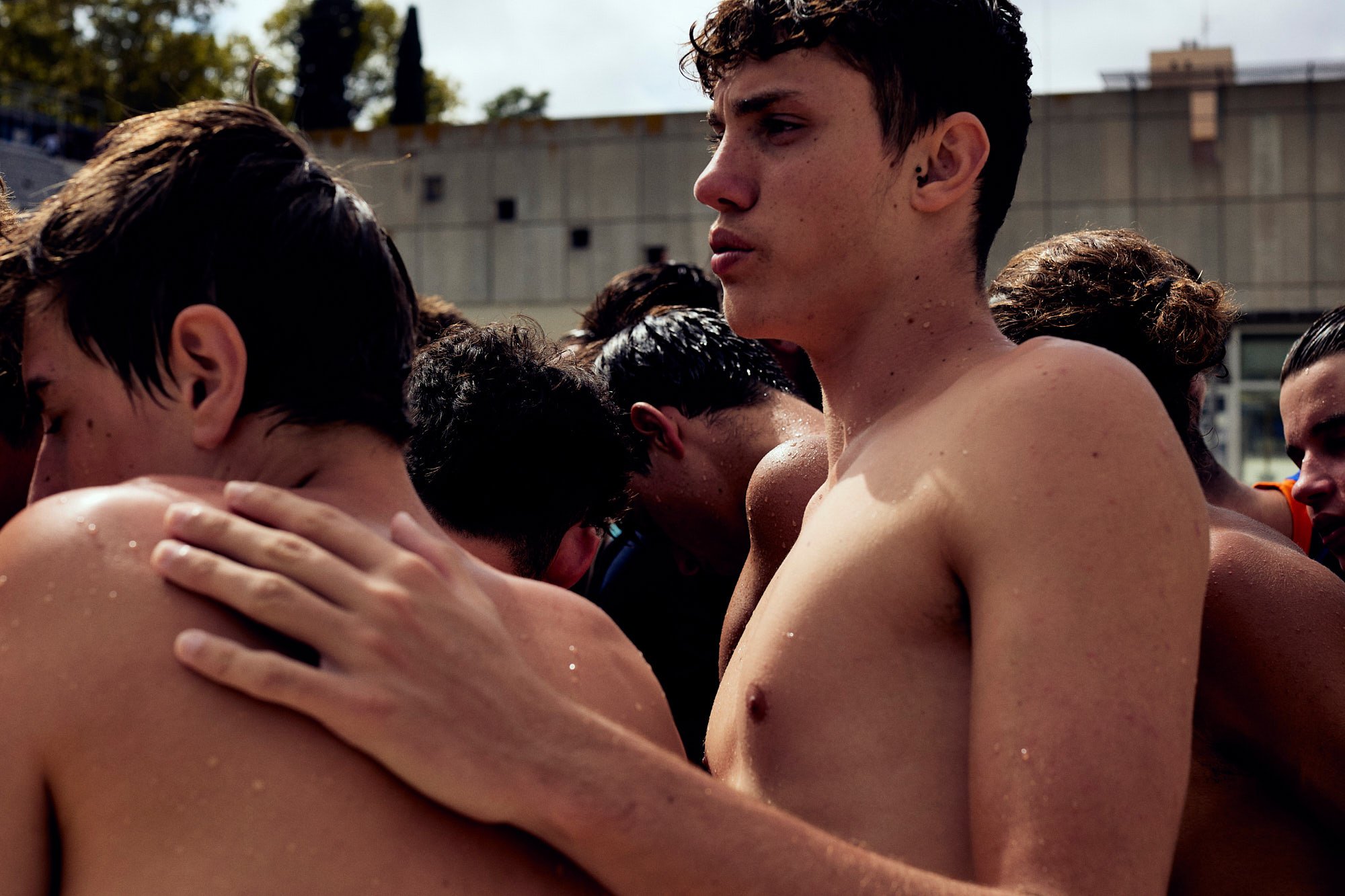 A group of young men shirtless, with wet skin, gathered close together outdoors, possibly before or after swimming or sports, with an overcast sky and building in the background.
