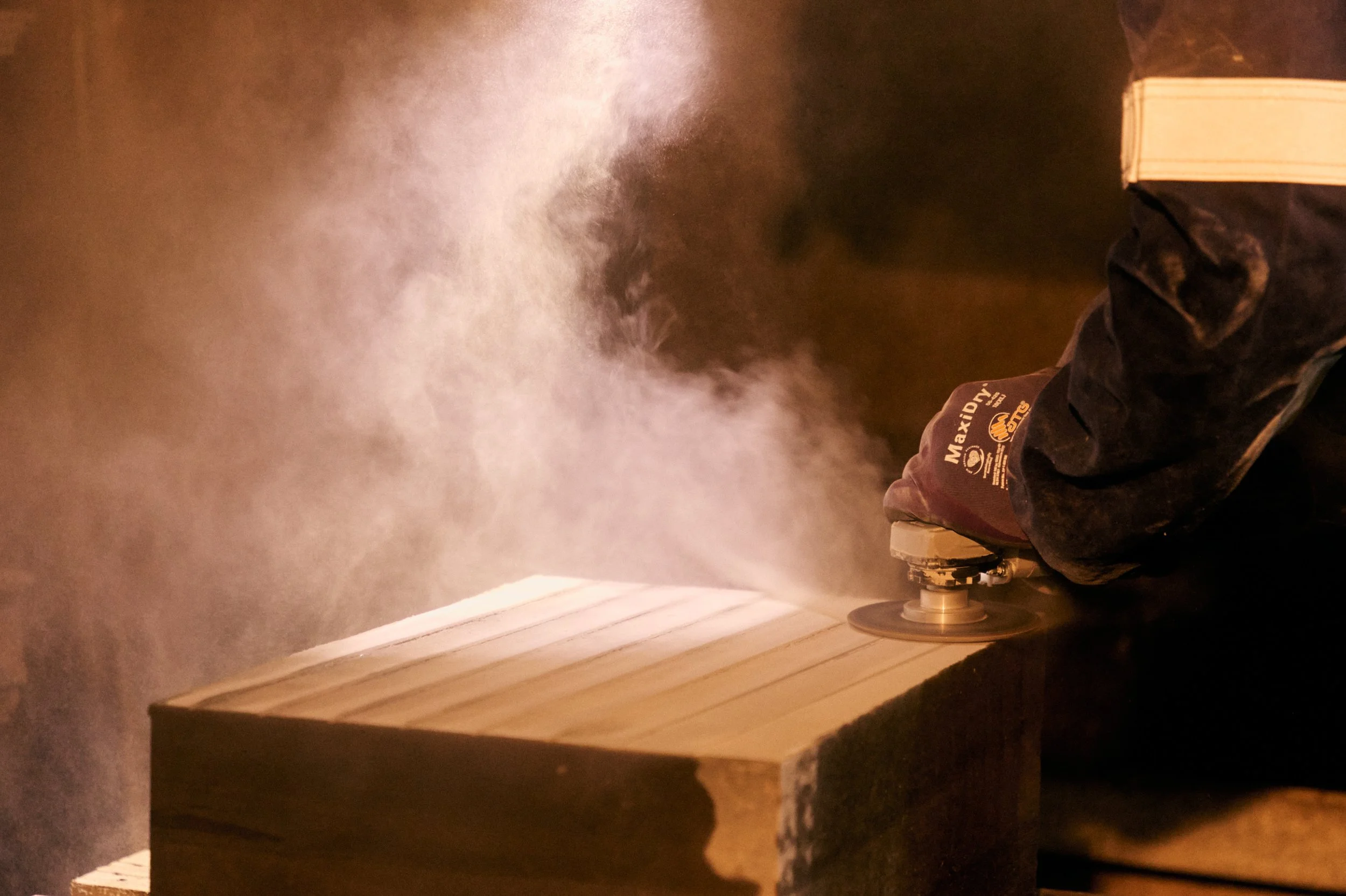 A person wearing gloves sanding a wooden surface with a handheld sander, producing a cloud of dust.