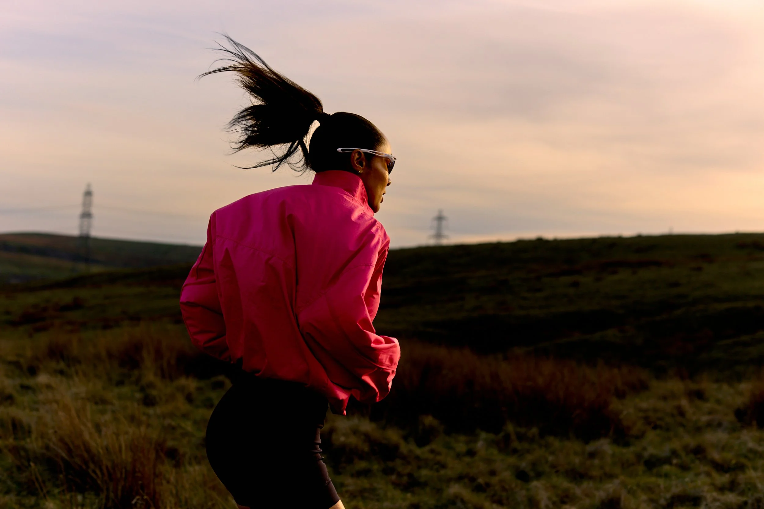Woman in a pink jacket running outdoors at sunset, with wind blowing her hair