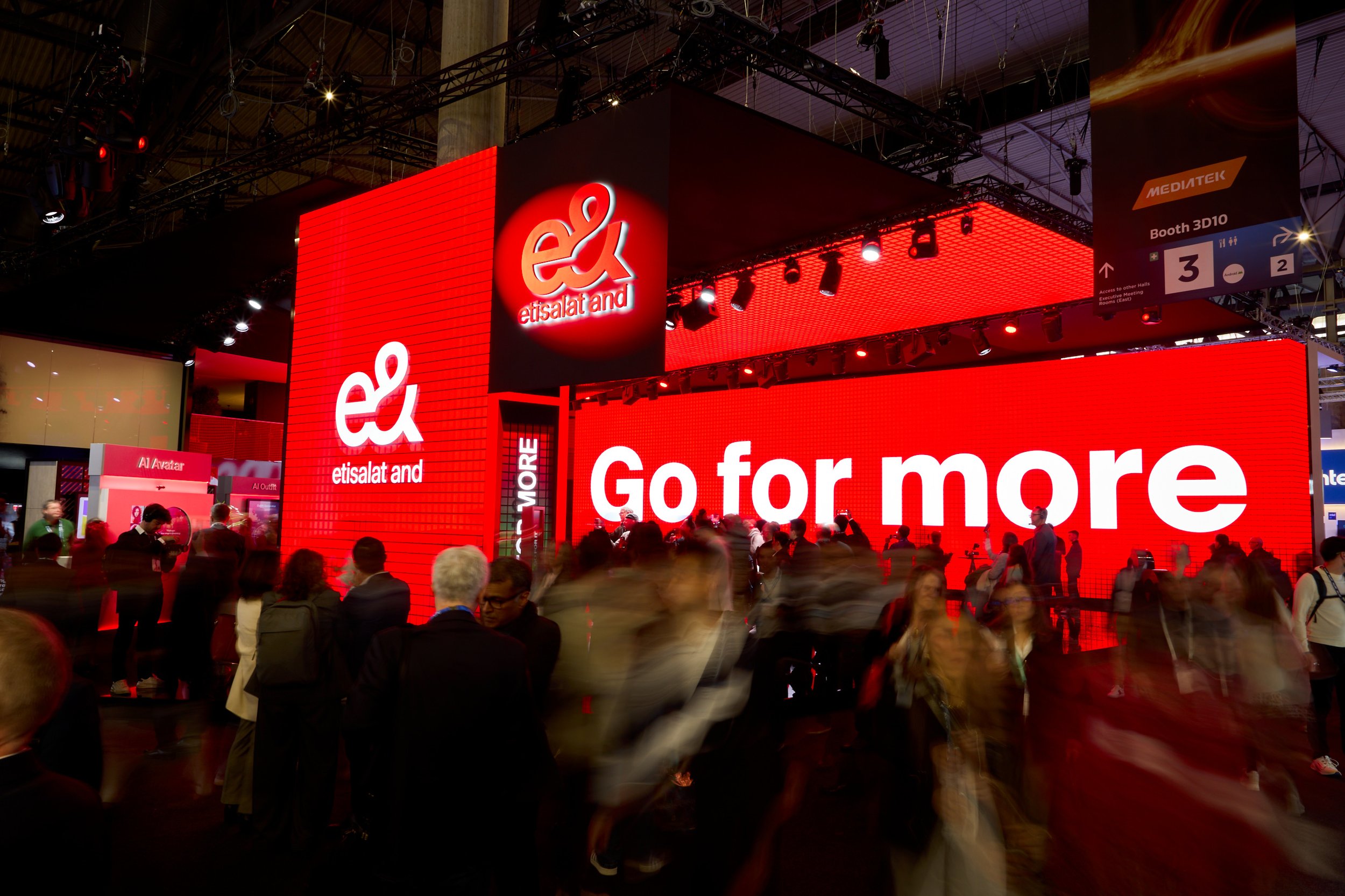 Trade show or conference booth for Etisalat and etisalat and featuring bright red digital advertising displays with the slogan "Go for more." Several people are walking and standing in front of the booth.