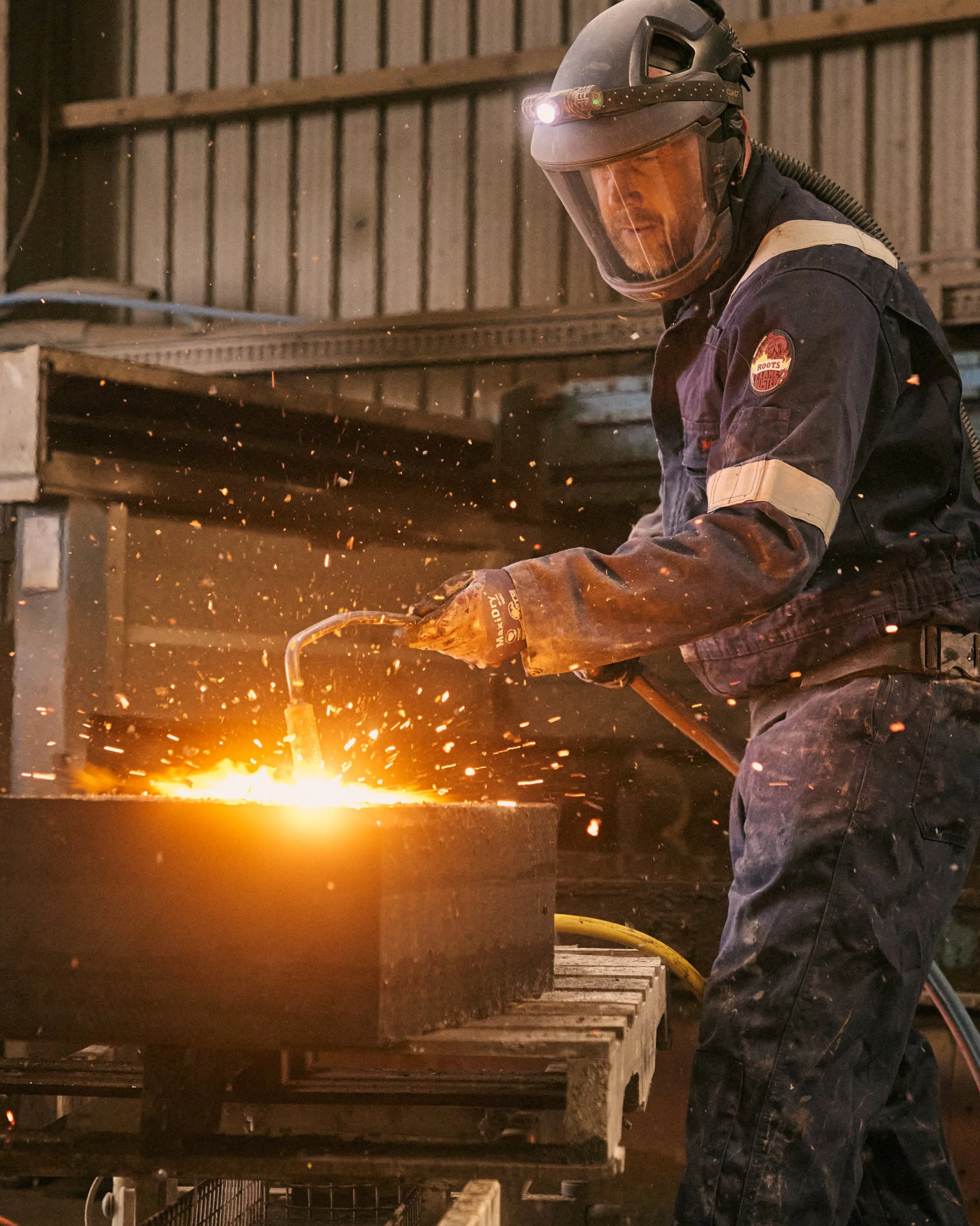 A worker wearing protective gear, including a helmet with a headlamp and gloves, is welding a large metal piece in an industrial workshop, with sparks flying from the welding process.