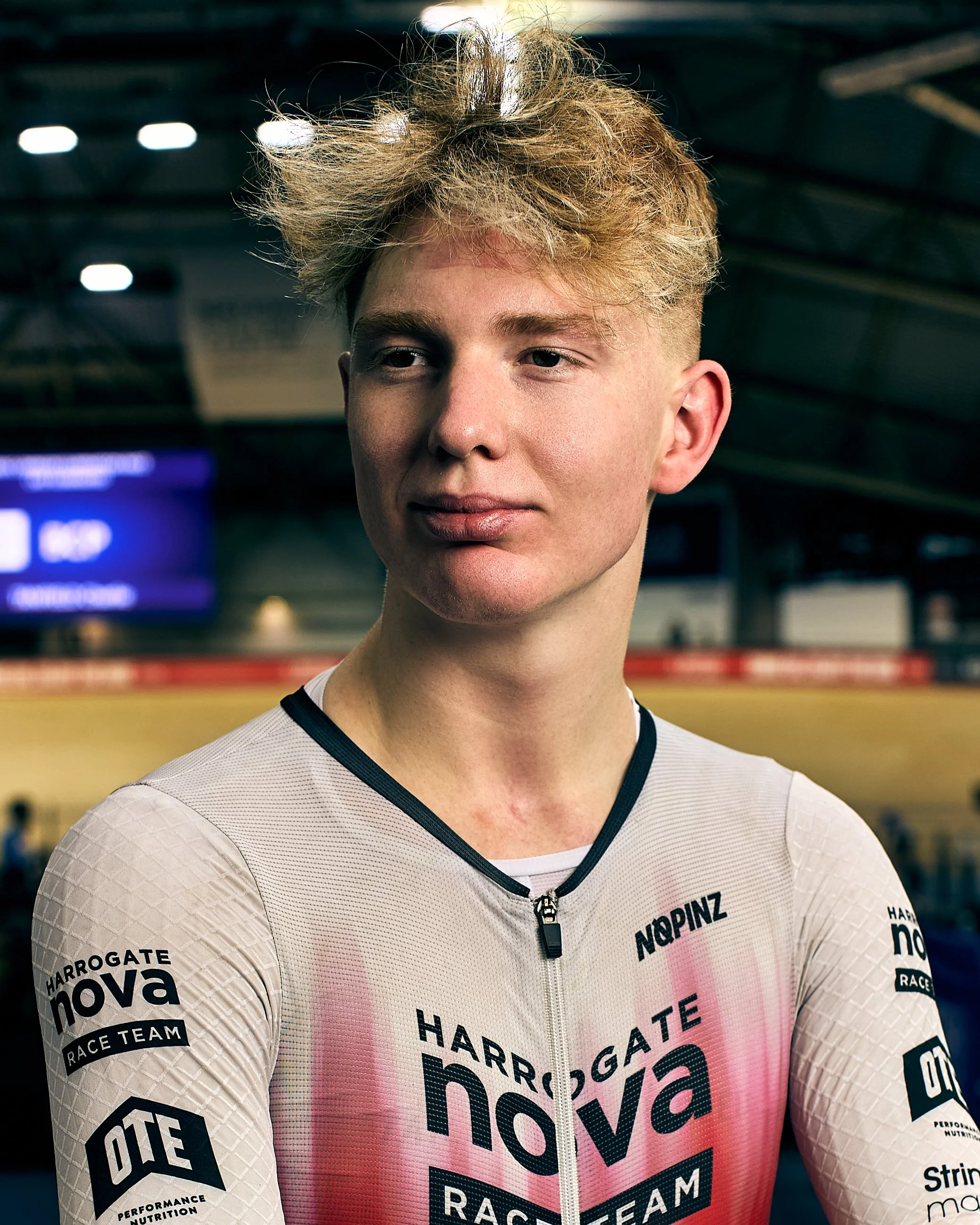 A young female cyclist with short, curly blonde hair wearing a white Harrrogate Nova Race Team jersey, standing inside an indoor velodrome.