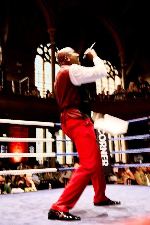 A man in red pants and a white shirt with a black vest, standing inside a boxing ring, holding a microphone and a white object, with a large audience and ornate windows in the background.