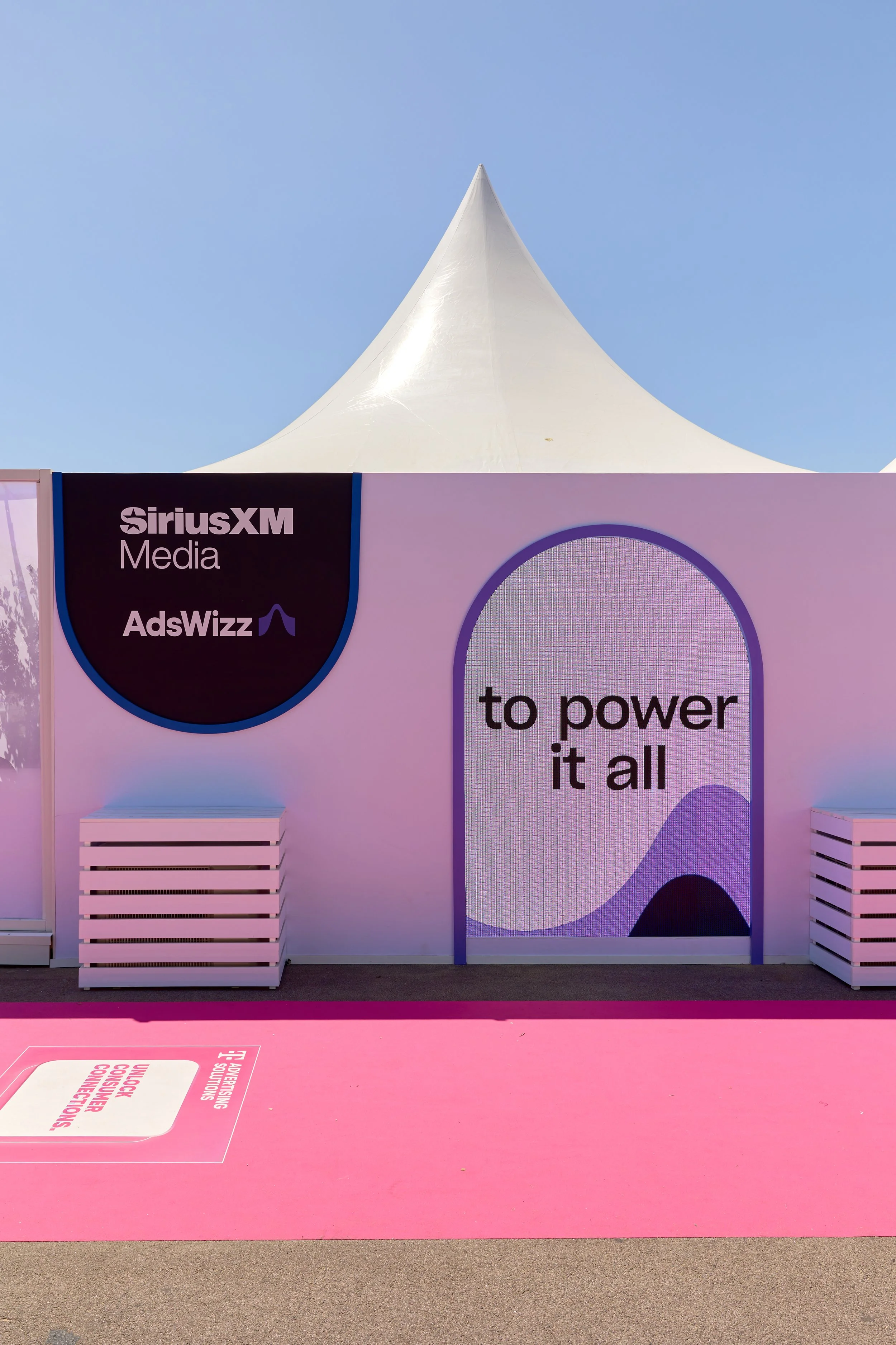A promotional event booth with SiriusXM Media and AdsWizz branding, featuring a large screen displaying the words 'to power it all' and an arch-shaped design, set against a blue sky.