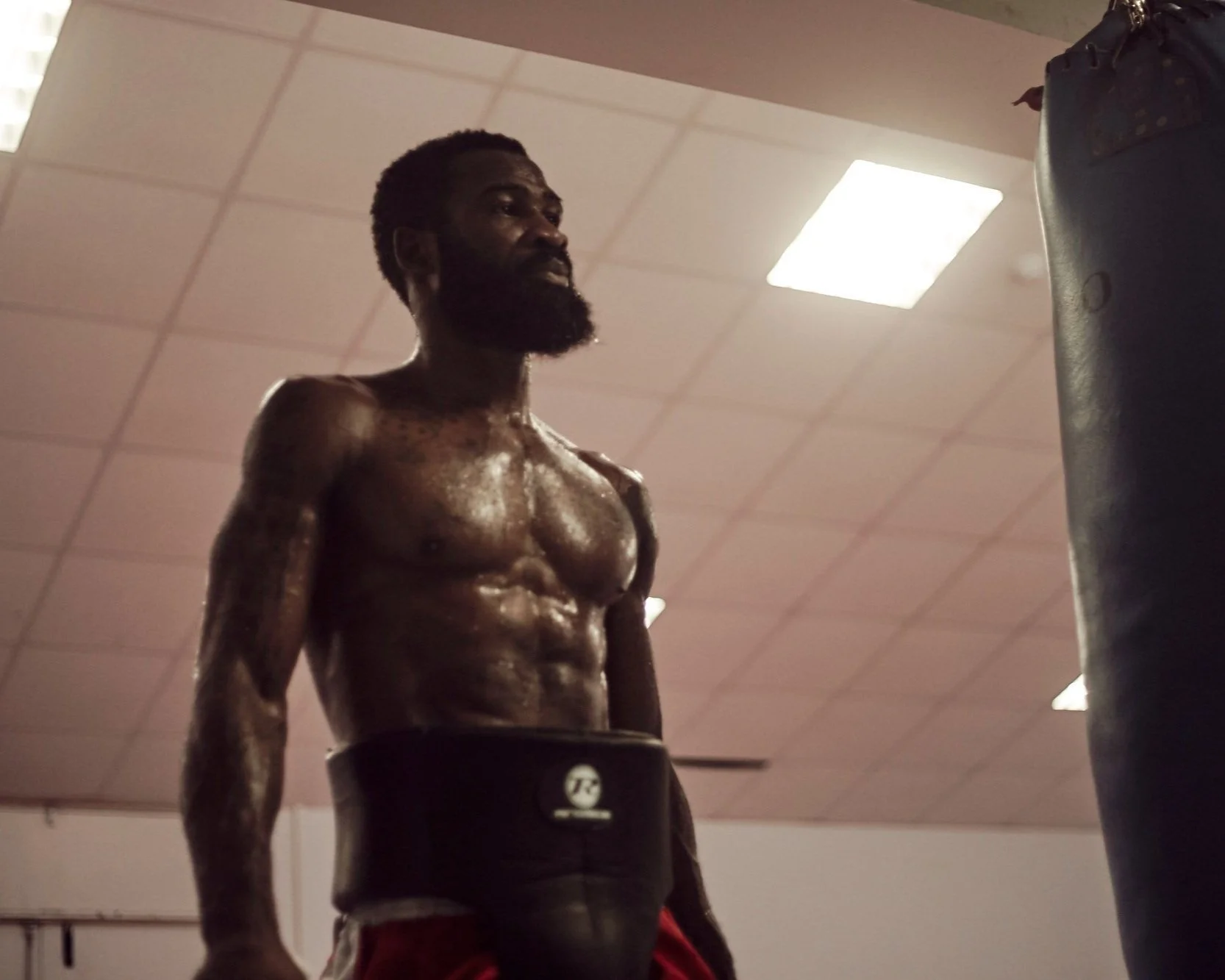 A shirtless man with a beard and short hair stands in a gym, focusing on a punching bag in front of him. His body is glistening with sweat under the gym lighting.