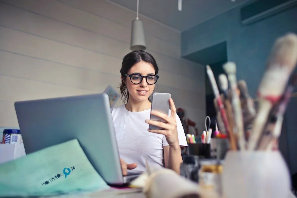 A woman with glasses looks at her phone while sitting at a desk with a laptop, paintbrushes, and art supplies.