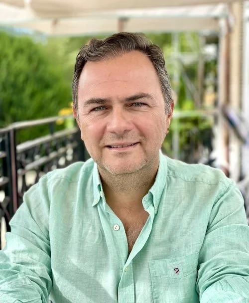 Baris Onay with short dark hair and a light green button-up shirt, smiling outdoors on a porch or balcony with green trees in the background.
