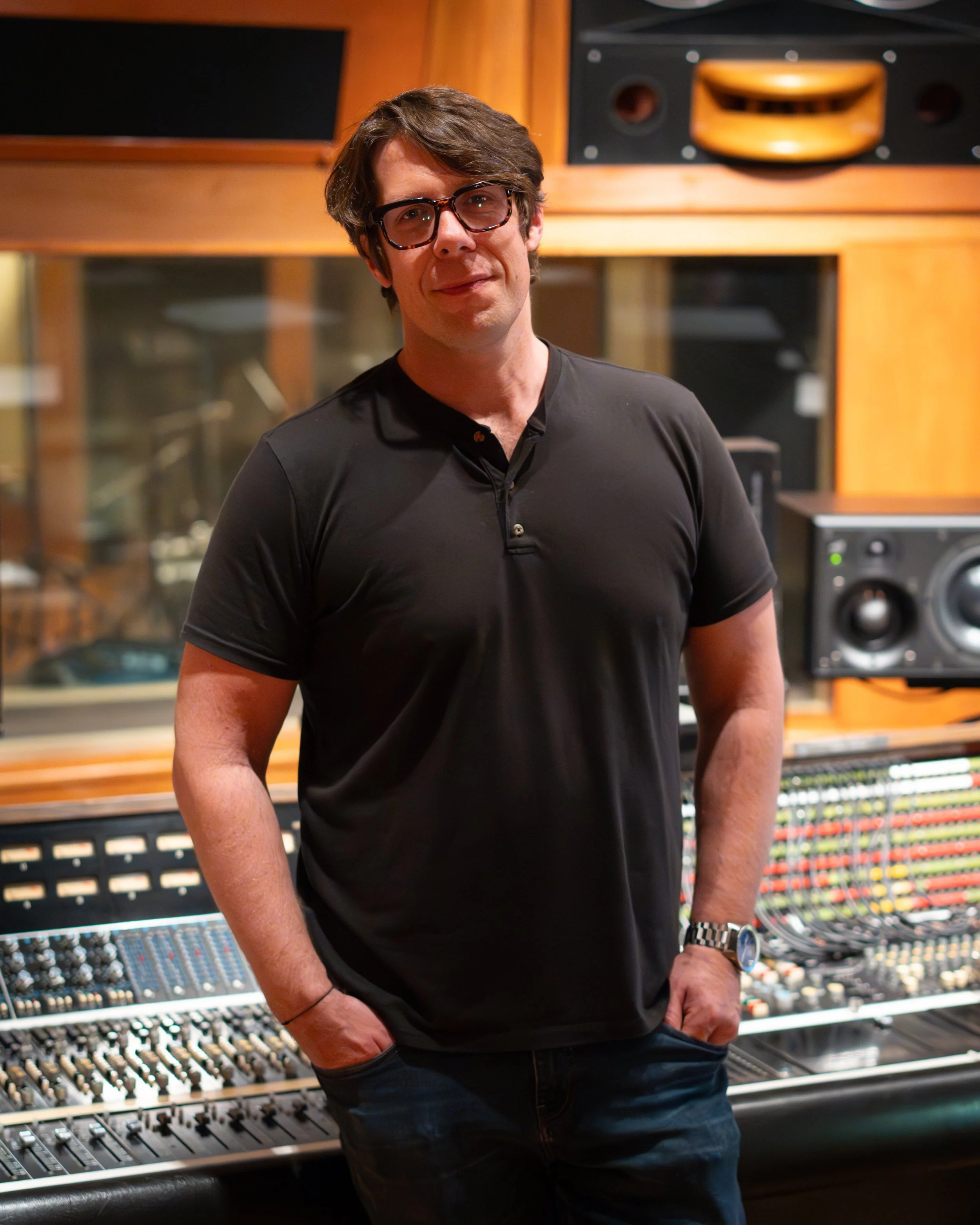 Photograph of recording engineer, standing in a recording studio control room, in front of a large format API recording console and a glass window that shows mic stands and instruments set up in the live room of the studio.