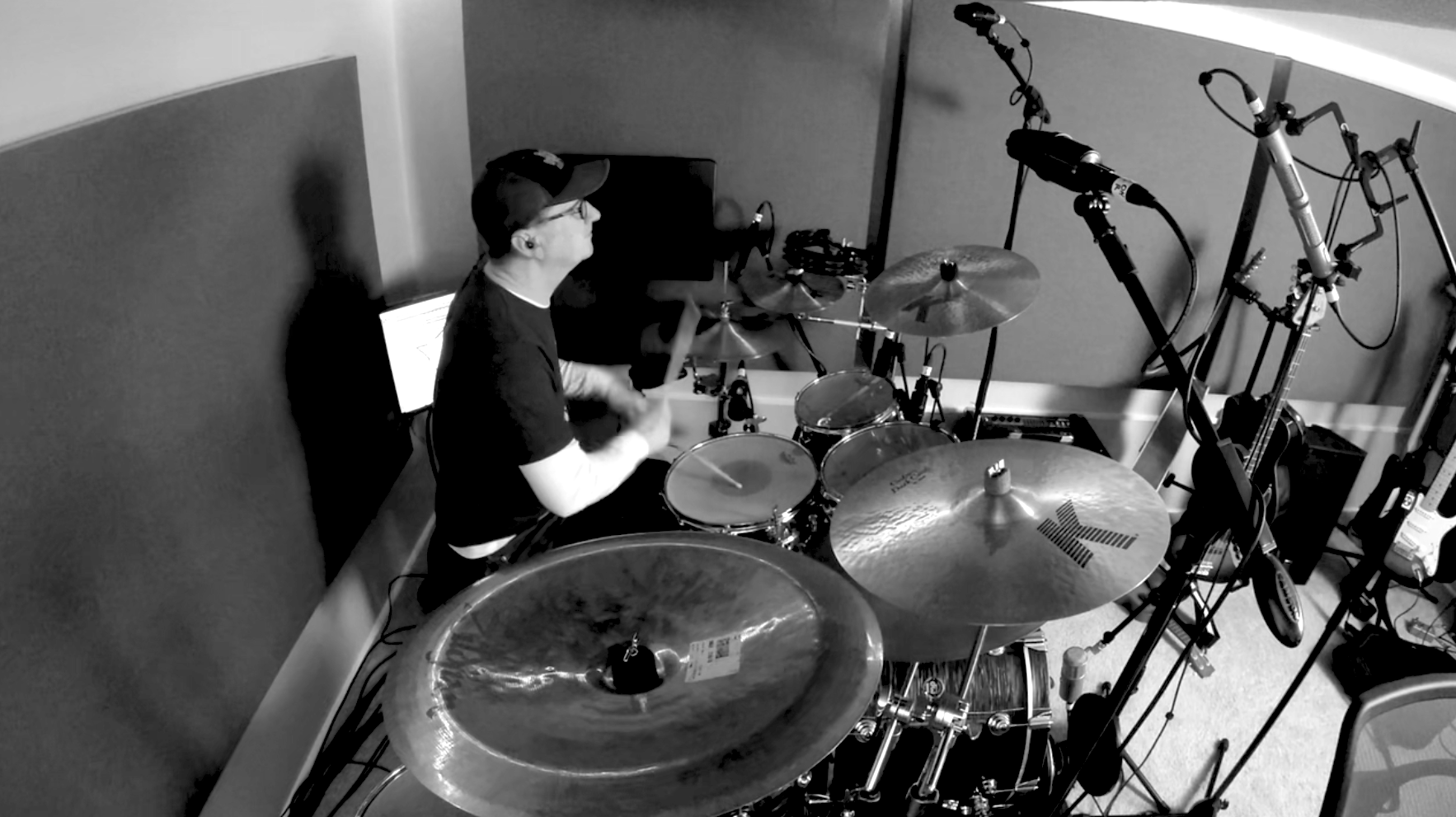 Man playing drum kit in a recording studio with acoustically treated walls, surrounded by microphones on mic stands, with a small computer monitor off to his left side.