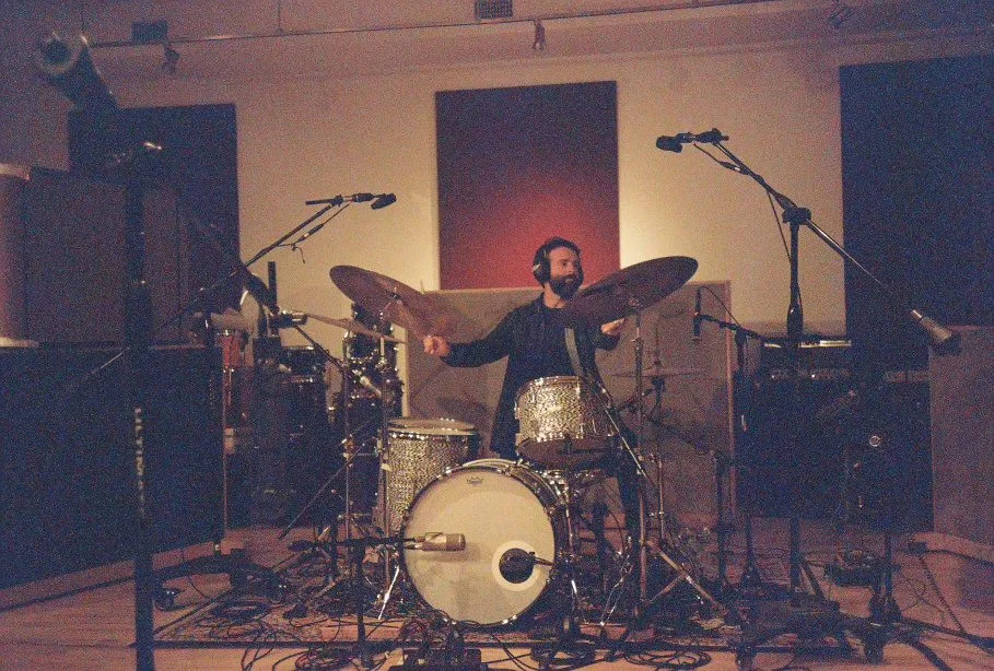 Drummer playing a drum kit in a dimly lit recording studio, surrounded by microphones, in front of a wall covered in acoustic treatment panels