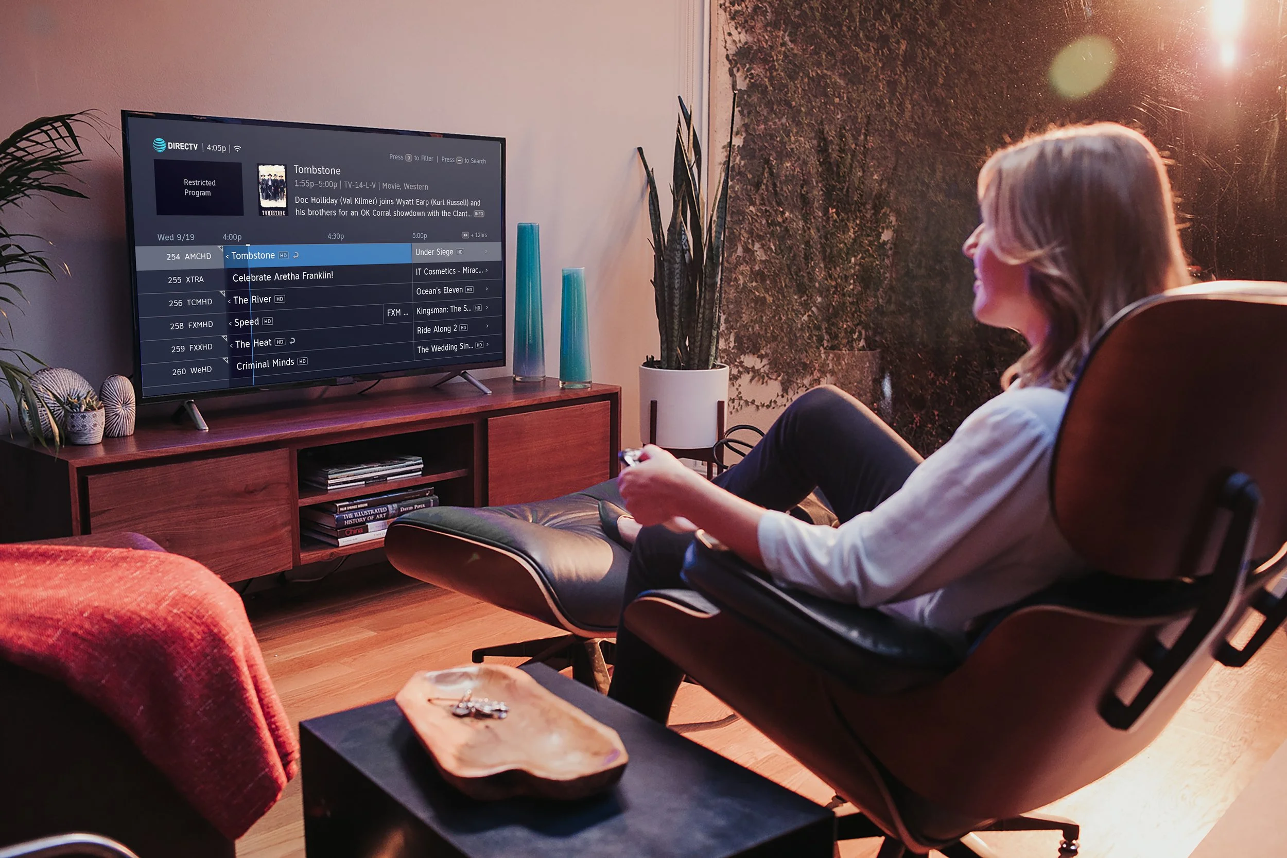 A woman sitting in a recliner watching television, holding a remote control. The room has a wooden TV stand with decorative vases and plants, a patterned wall or window shade behind her, and a small table with a wooden tray in front of her.