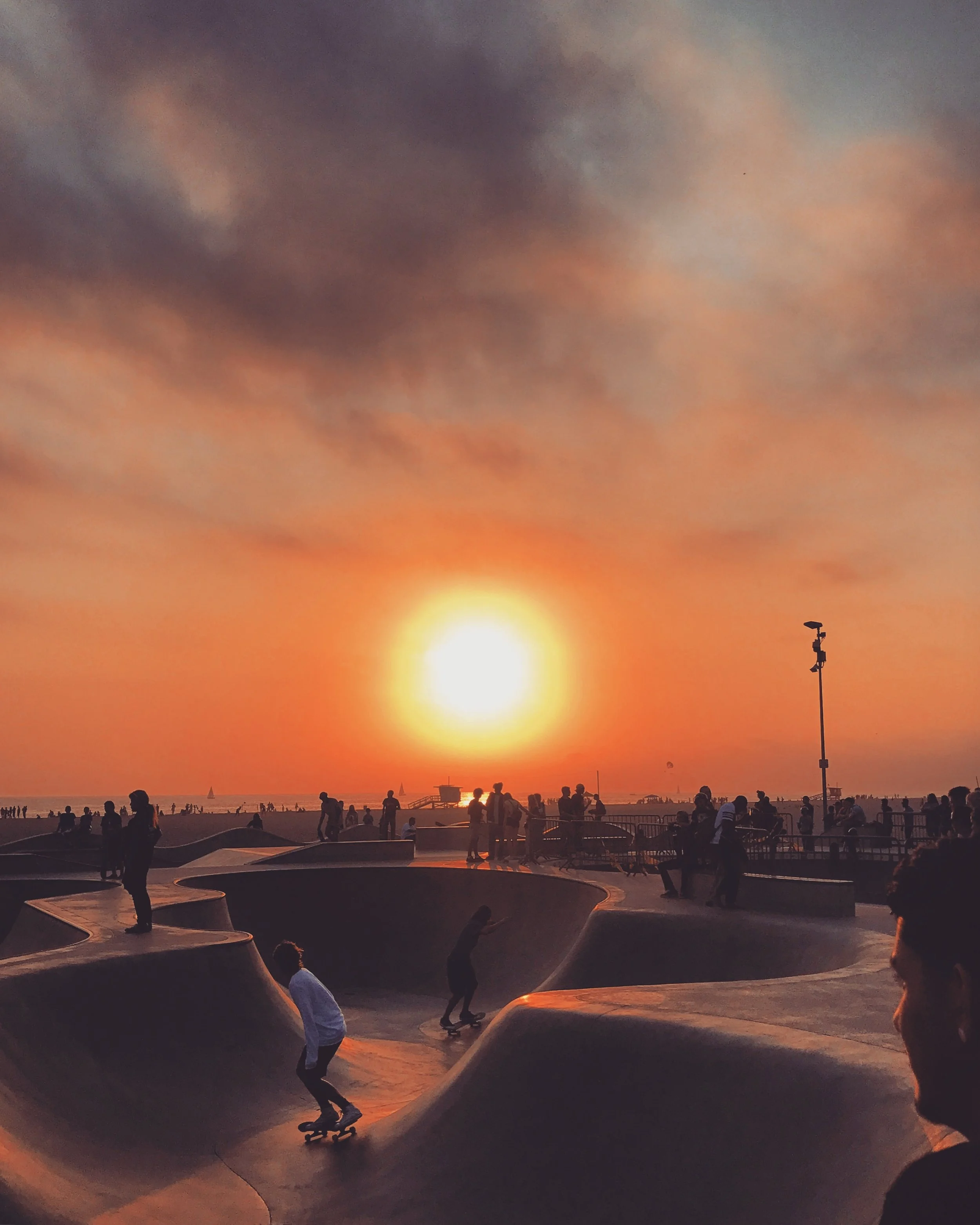 Skateboarders and people at a skate park during sunset with a cloudy sky and a lighthouse in the background.