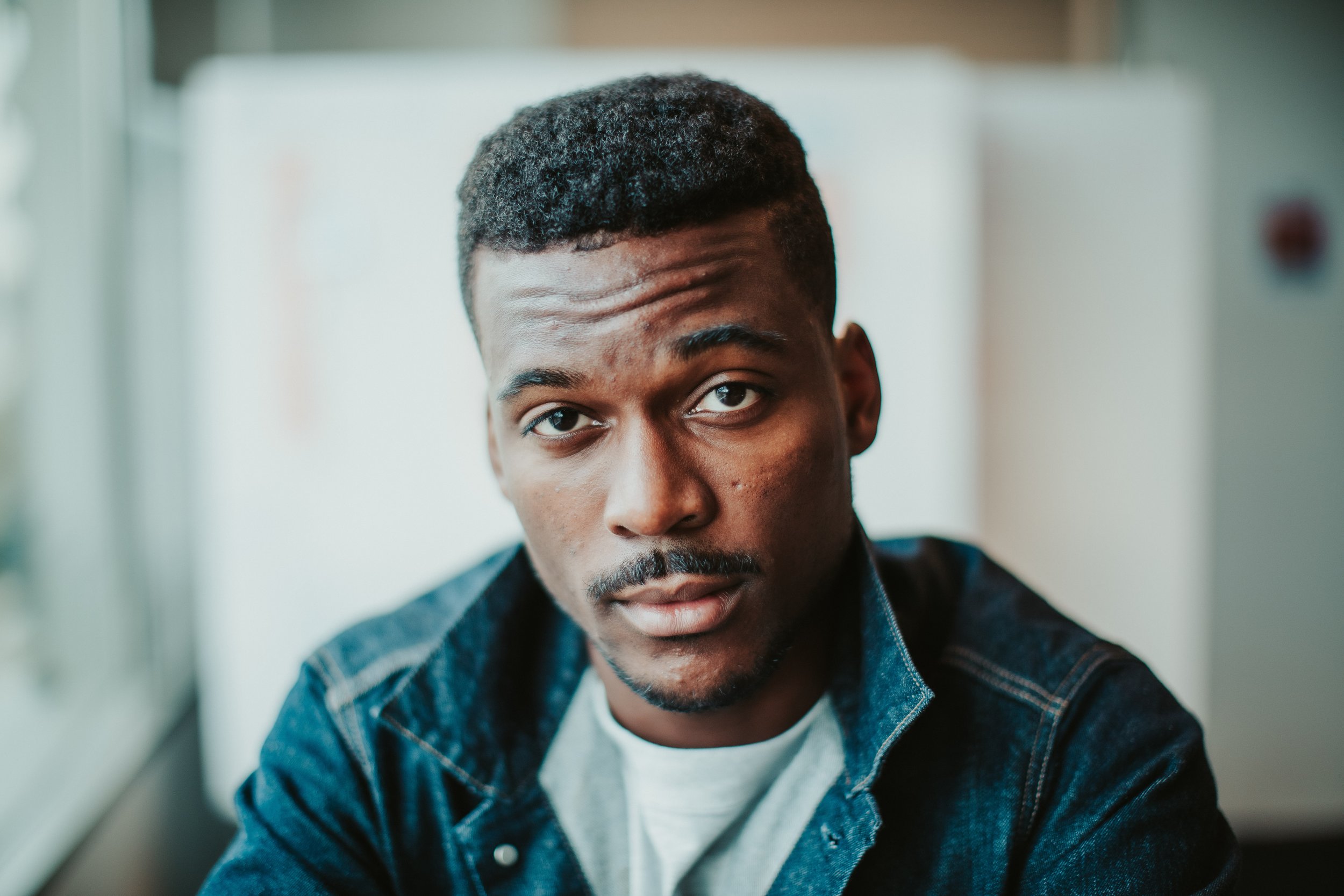Close-up of a young man with dark hair, wearing a denim jacket, looking directly at the camera with a serious expression, in a softly lit room.