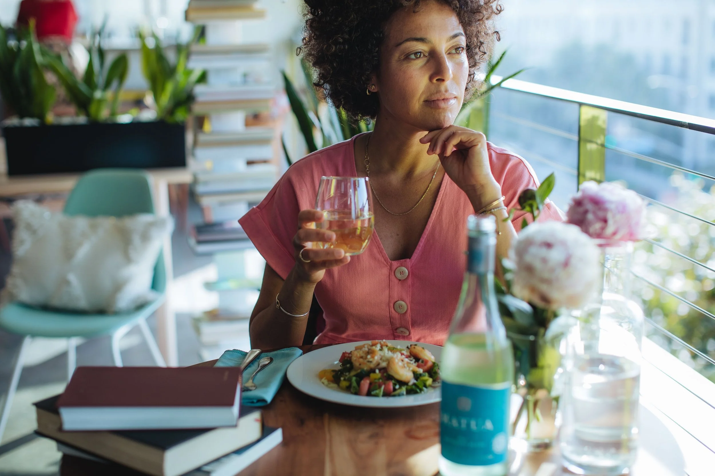 A woman with curly hair sitting at a table in a restaurant, holding a glass of white wine, with a plate of salad in front of her, and sunlight coming through the window.