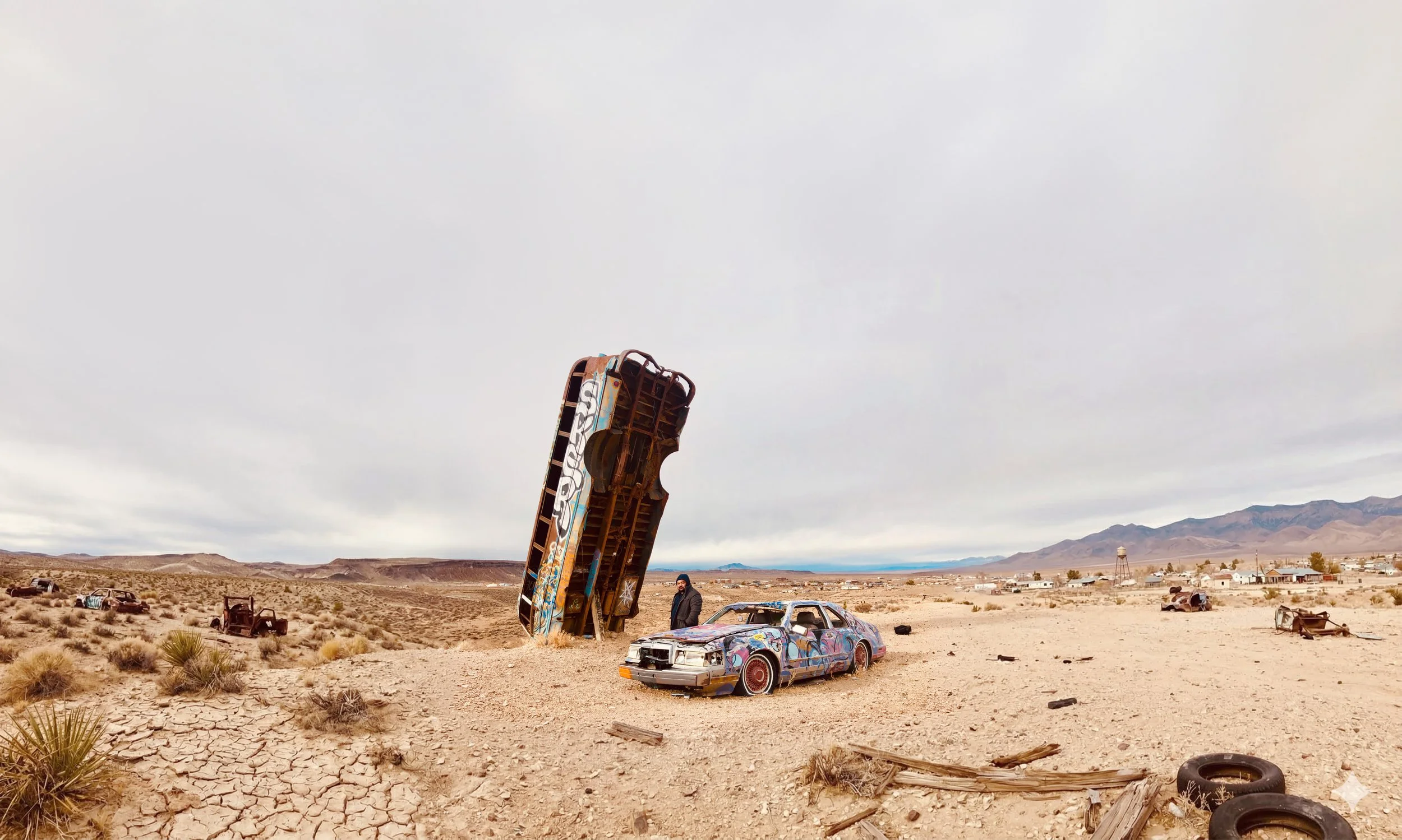 Desert landscape with abandoned, graffiti-covered cars, an upright large rusted metal structure, and mountains in the background. Overcast sky.