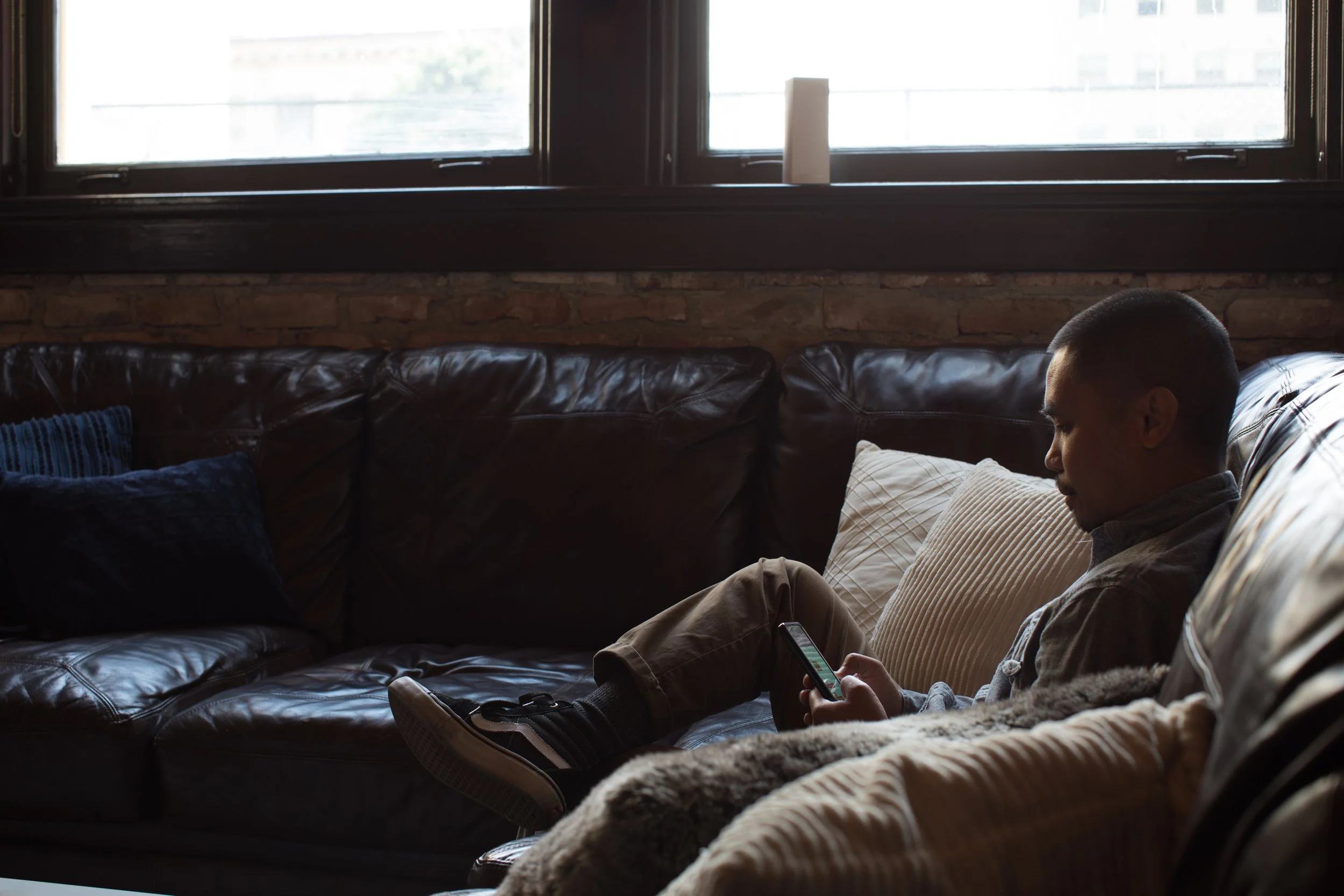 Young man sitting on a leather couch, looking at a smartphone, with pillows behind him and a window above.