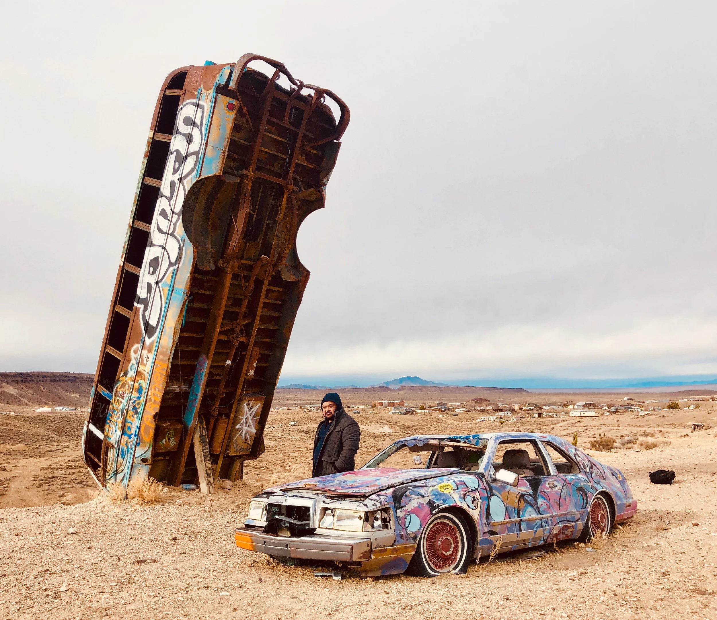 A man standing on desert terrain next to a colorful, graffiti-covered abandoned car and a rusty, upside-down, graffiti-covered airplane fuselage in a barren landscape with distant mountains and overcast sky.