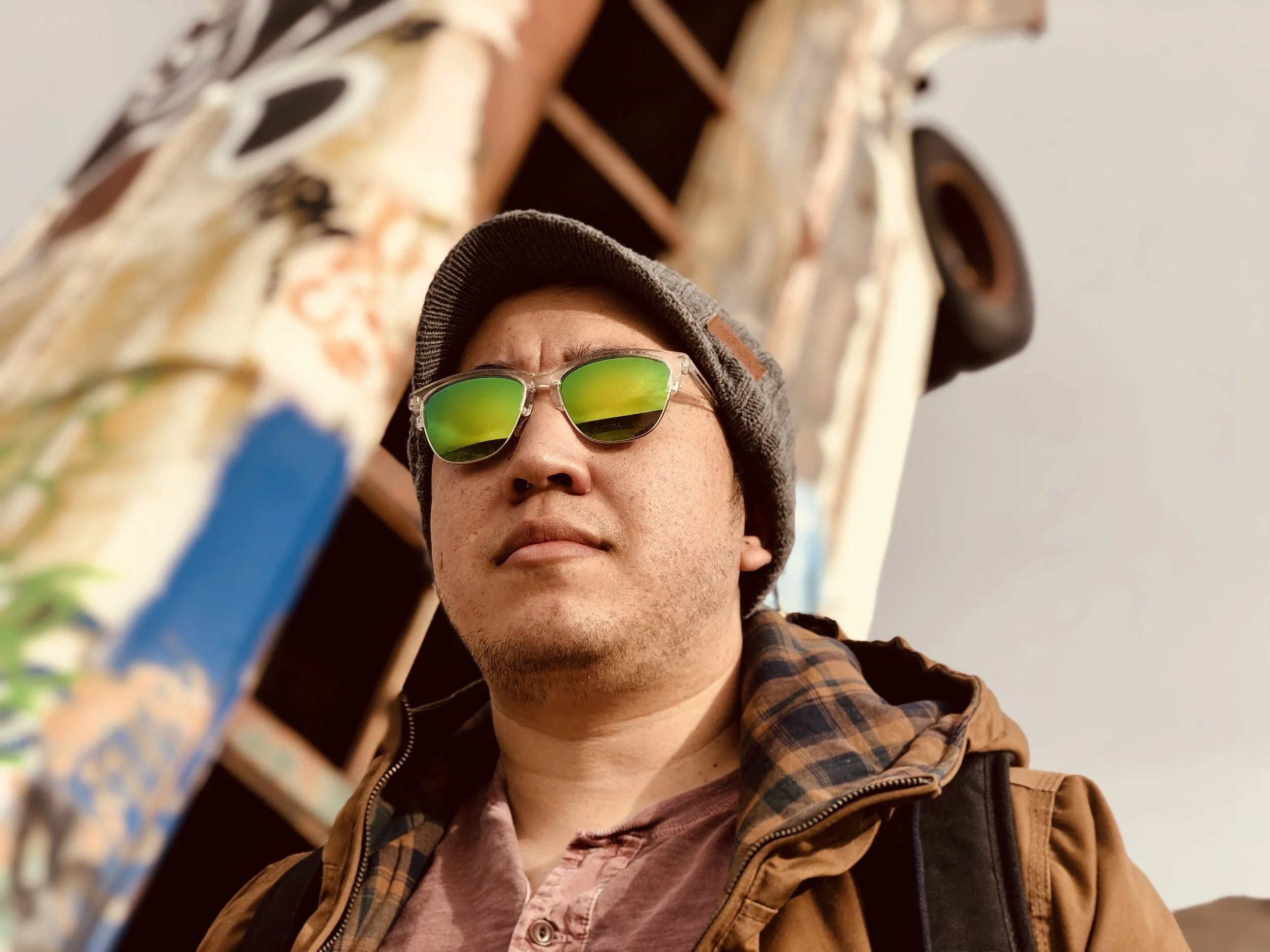 A man wearing reflective green sunglasses, a brown jacket, and a gray beanie, standing outdoors near a graffiti-covered structure with a tire attached to it.