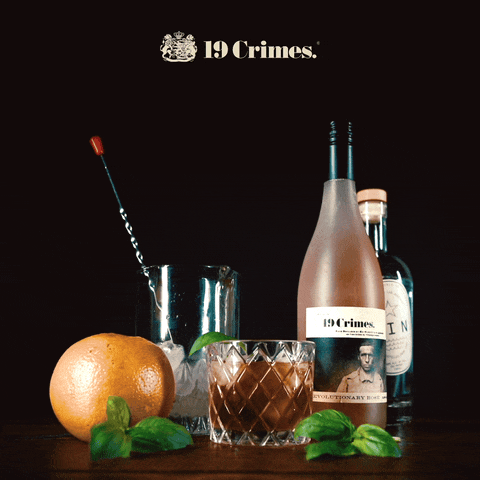 Antique bottles, a glass of cocktail, a grapefruit, mint leaves, and a cocktail stirrer on a dark surface