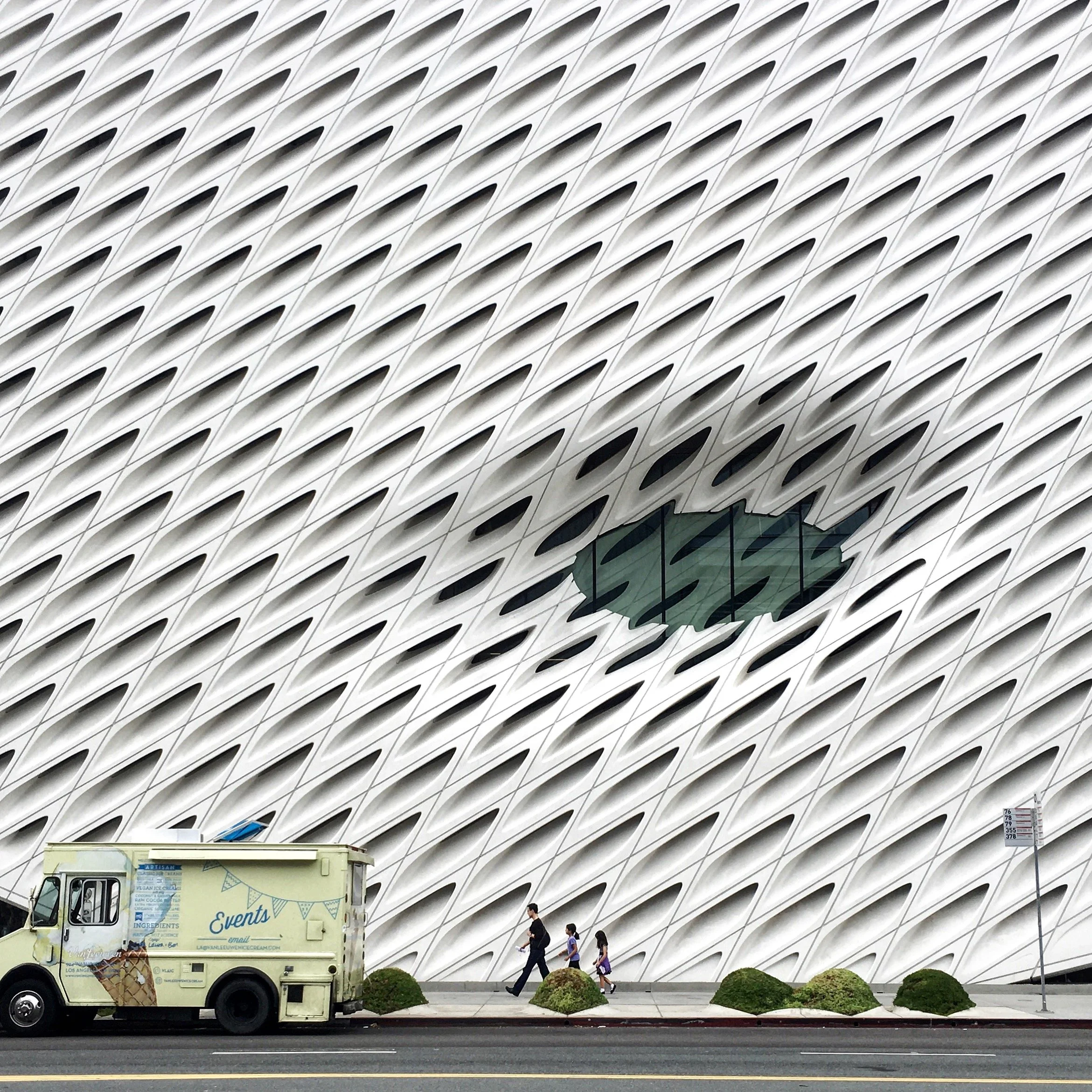 Modern building with a decorative white lattice facade, featuring a green oval window, and a small group of people walking past a food truck parked on the street.