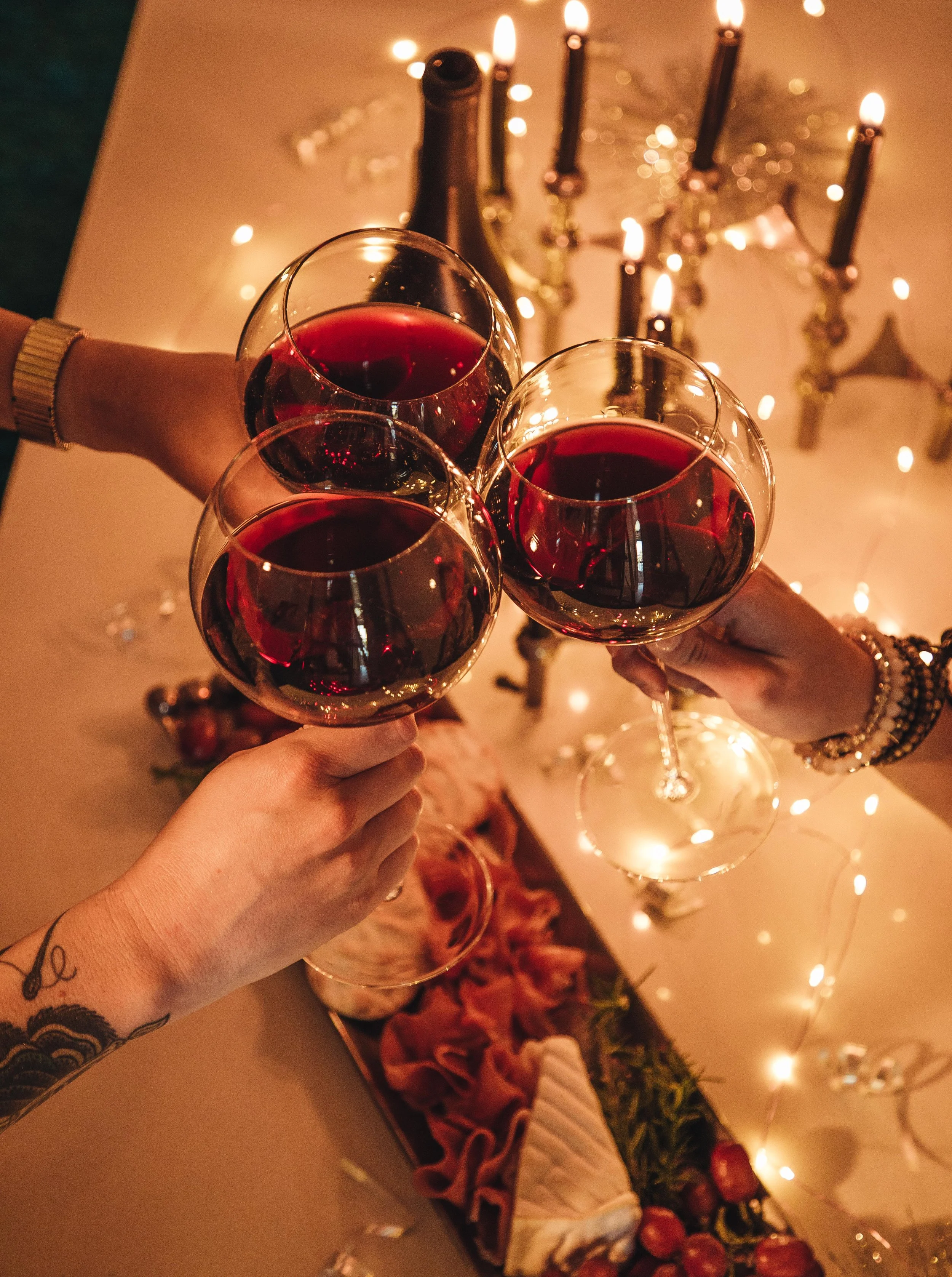 Three people clinking glasses of red wine during a celebration with a decorated table, candles, and fairy lights in the background.