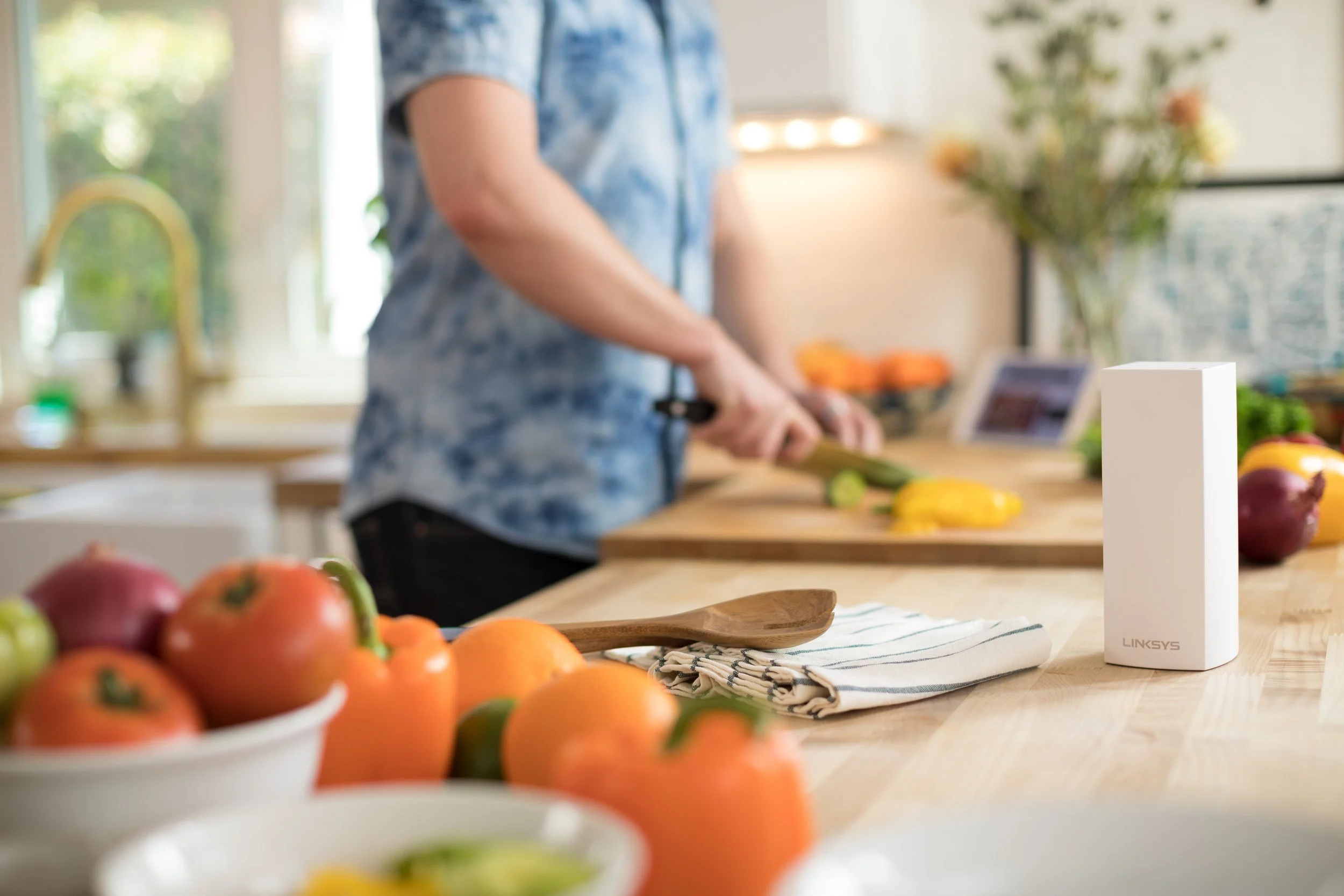 Person chopping vegetables on a kitchen countertop with fresh produce and a white Linksys device in the foreground.