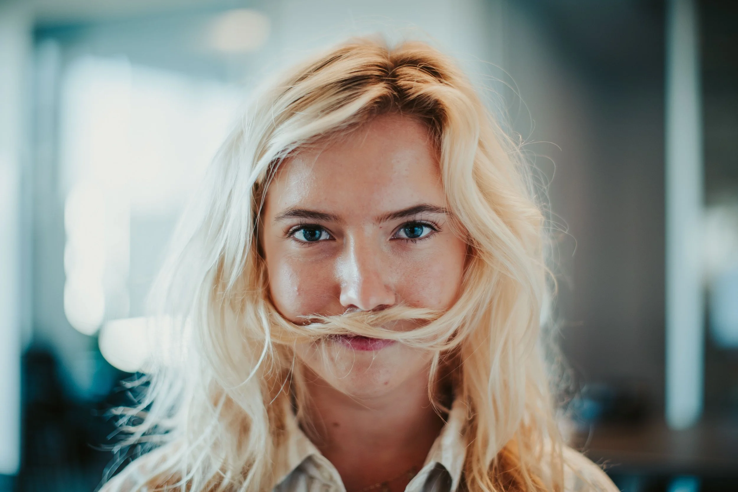 Close-up of a blonde woman with blue eyes, looking directly at the camera with a playful smile, in an indoor setting.