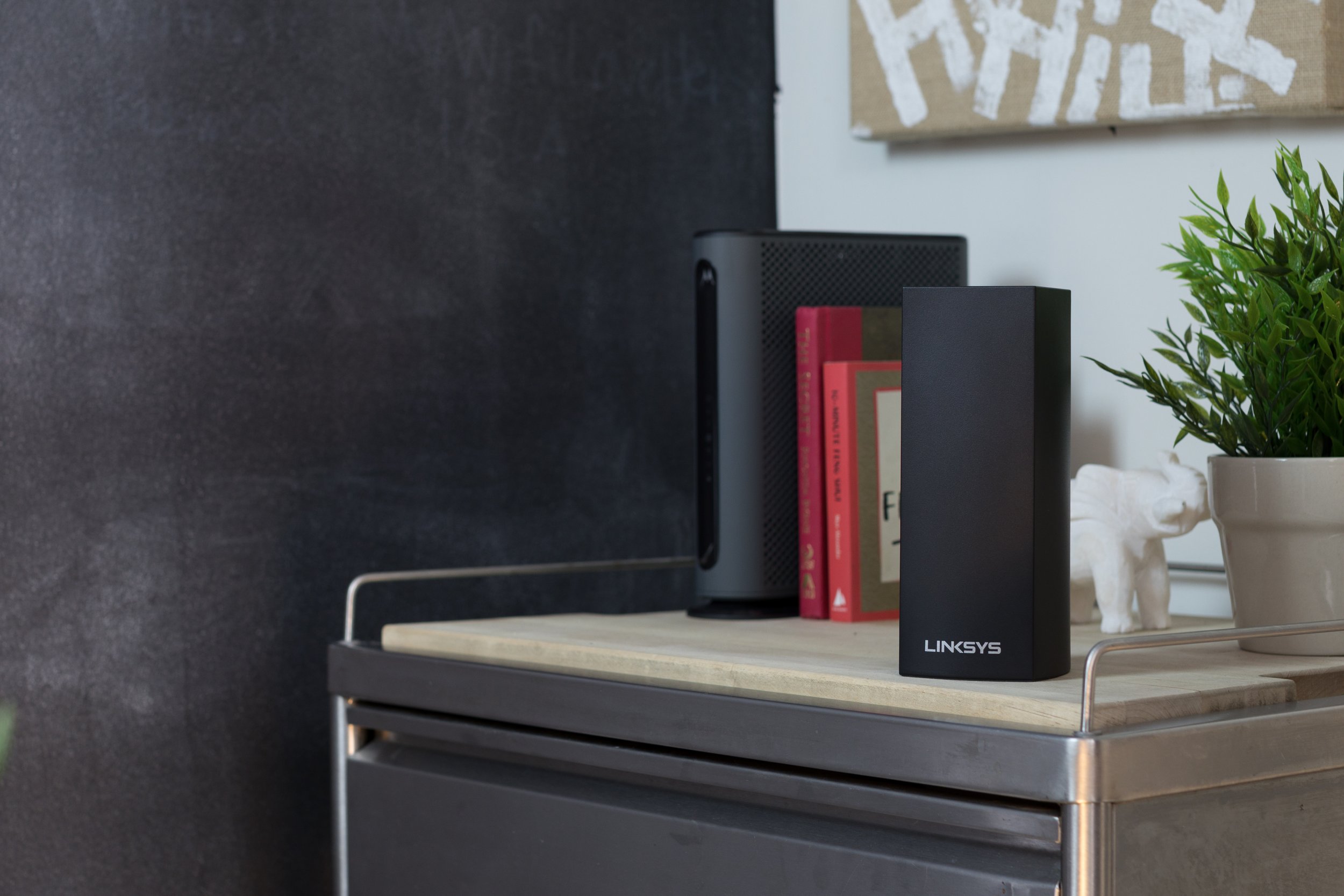 A black Linksys mesh Wi-Fi router on a wooden tray, with a small potted plant, a white decorative elephant, and some books in the background.