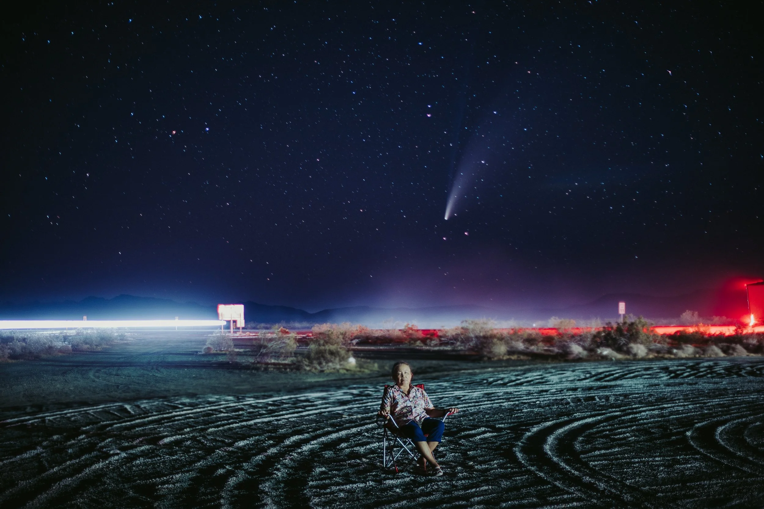 A woman sitting in a foldable chair on a desert dirt road at night, watching a comet and a starry sky with streaks of light from passing cars in the distance.