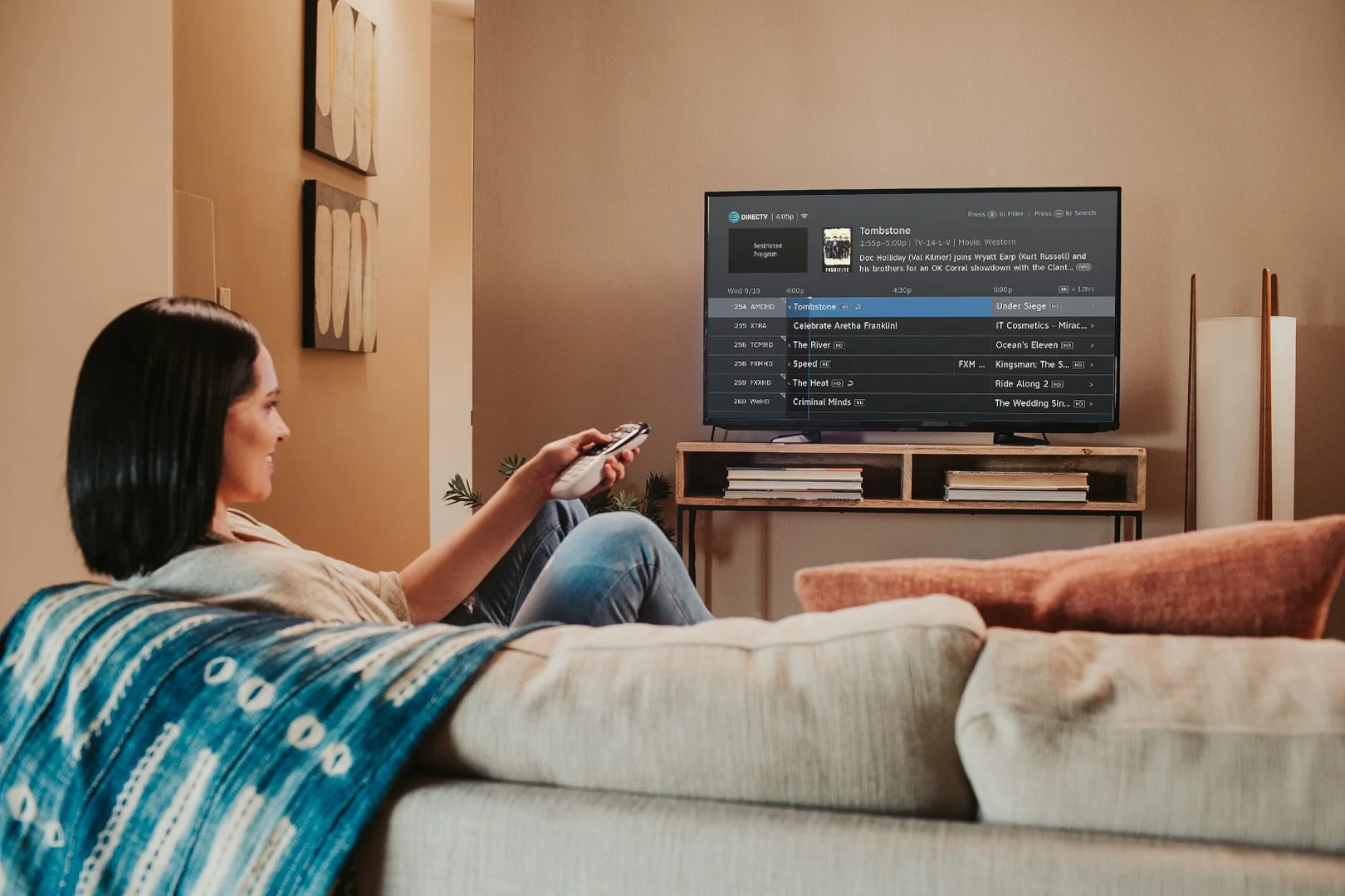 A woman with dark hair sitting on a beige couch in a living room, holding a remote control, watching TV. The TV displays a guide with multiple channels and shows. There are decorative art pieces on the wall and a plant on a stand beside the TV.