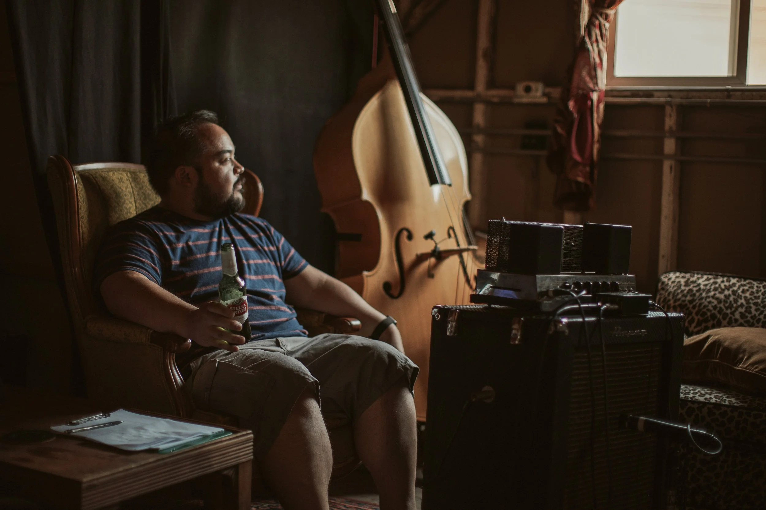 A man with a beard sitting on an armchair, holding a bottle of beer, in a dimly lit room with musical instruments including a double bass and a guitar, and electronic equipment on a table.