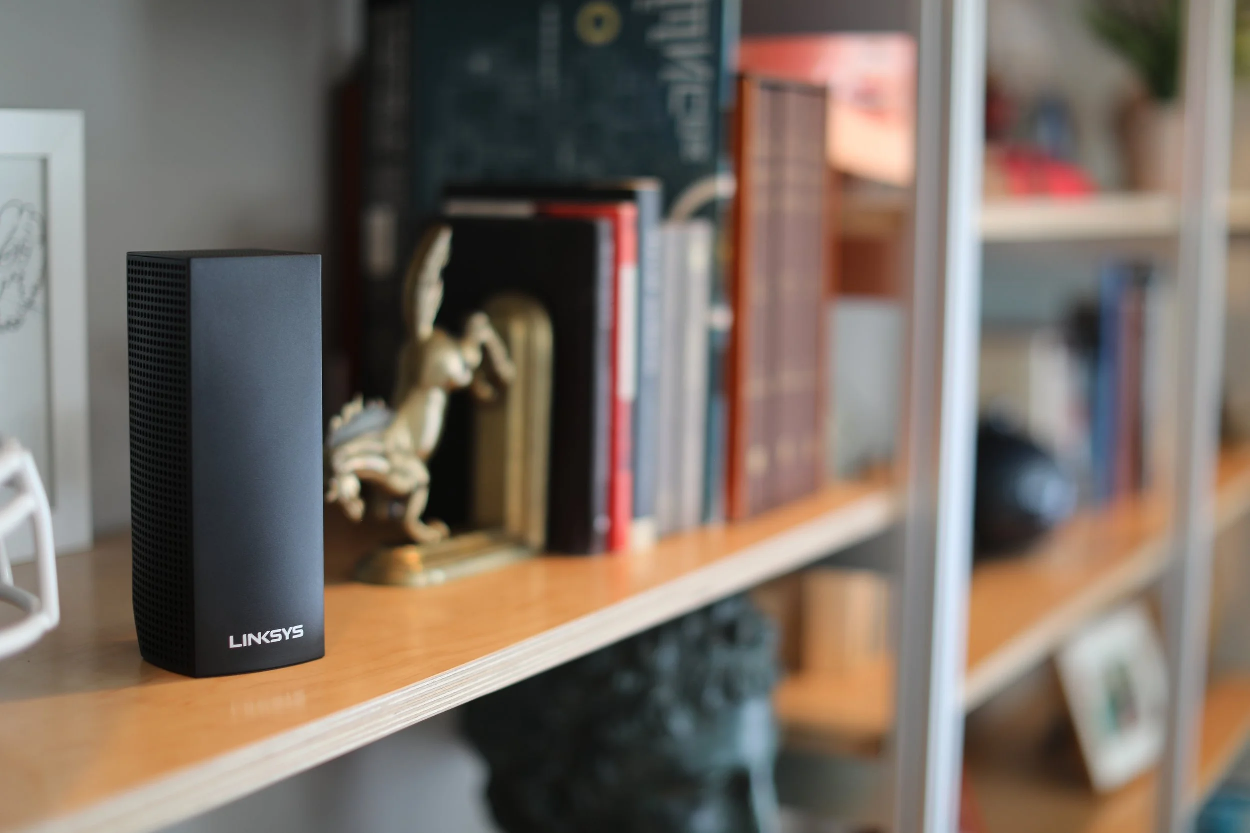 A black Linksys Wi-Fi router on a wooden bookshelf with books and decorative items in the background.