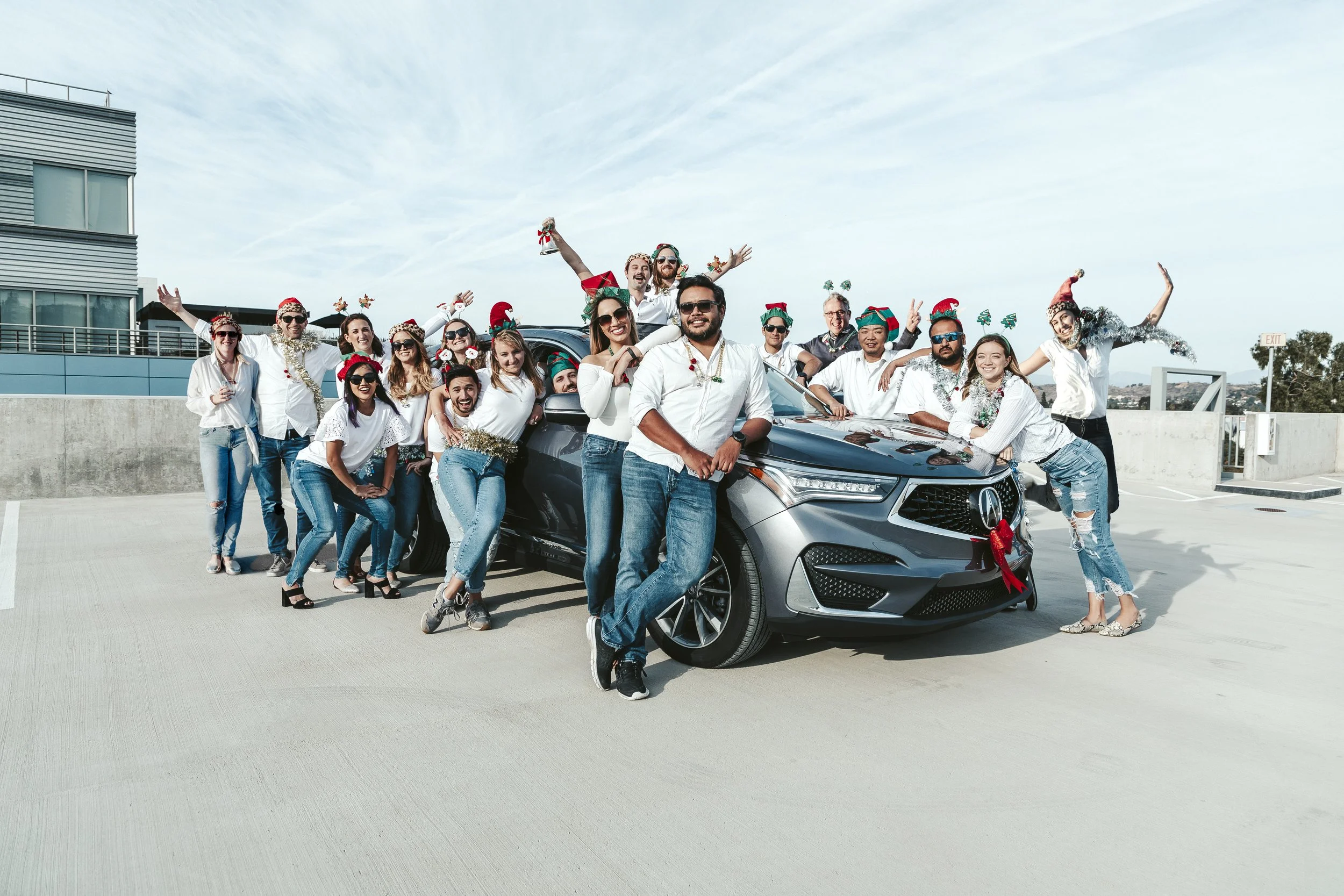 Group of people celebrating Christmas outdoors around a car, wearing festive hats and accessories.