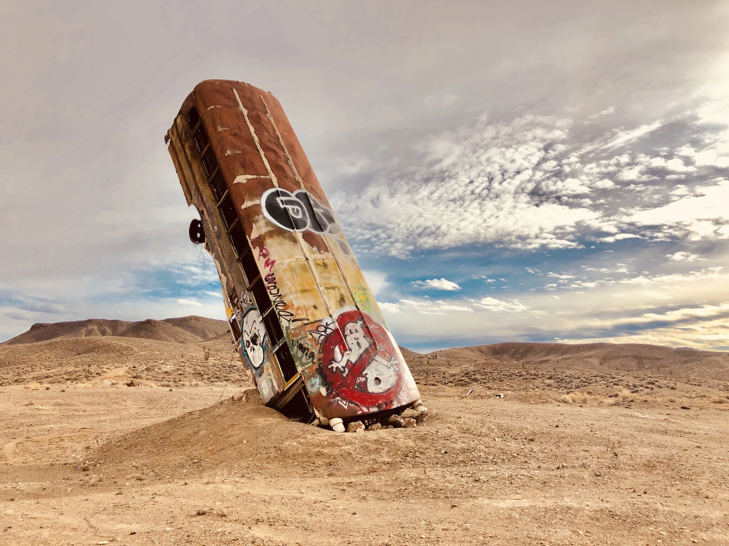An abandoned, graffiti-covered bus tilted on its side in a desert landscape under a cloudy sky.