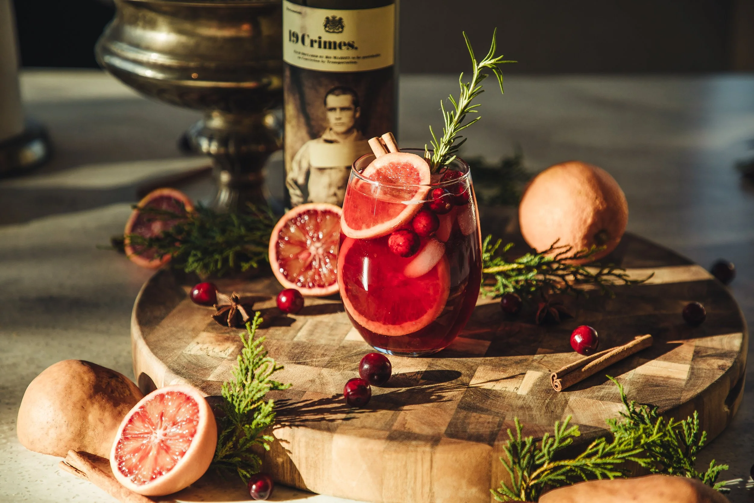 A glass of red beverage garnished with sliced grapefruit, cranberries, a sprig of rosemary, and cinnamon on a wooden board, surrounded by whole grapefruit, cranberries, cinnamon, and sprigs of greenery.