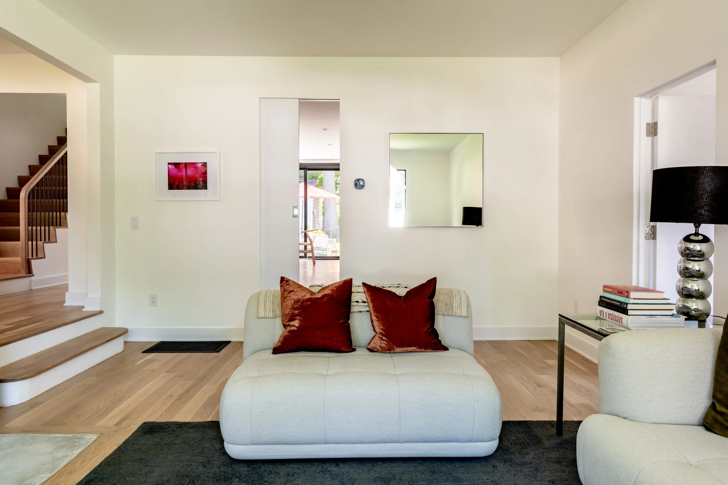 Living room with beige sofa, red pillows, black lamp, side table with books, wall mirror, artwork, wooden stairs visible to the left, and opens to a patio area.