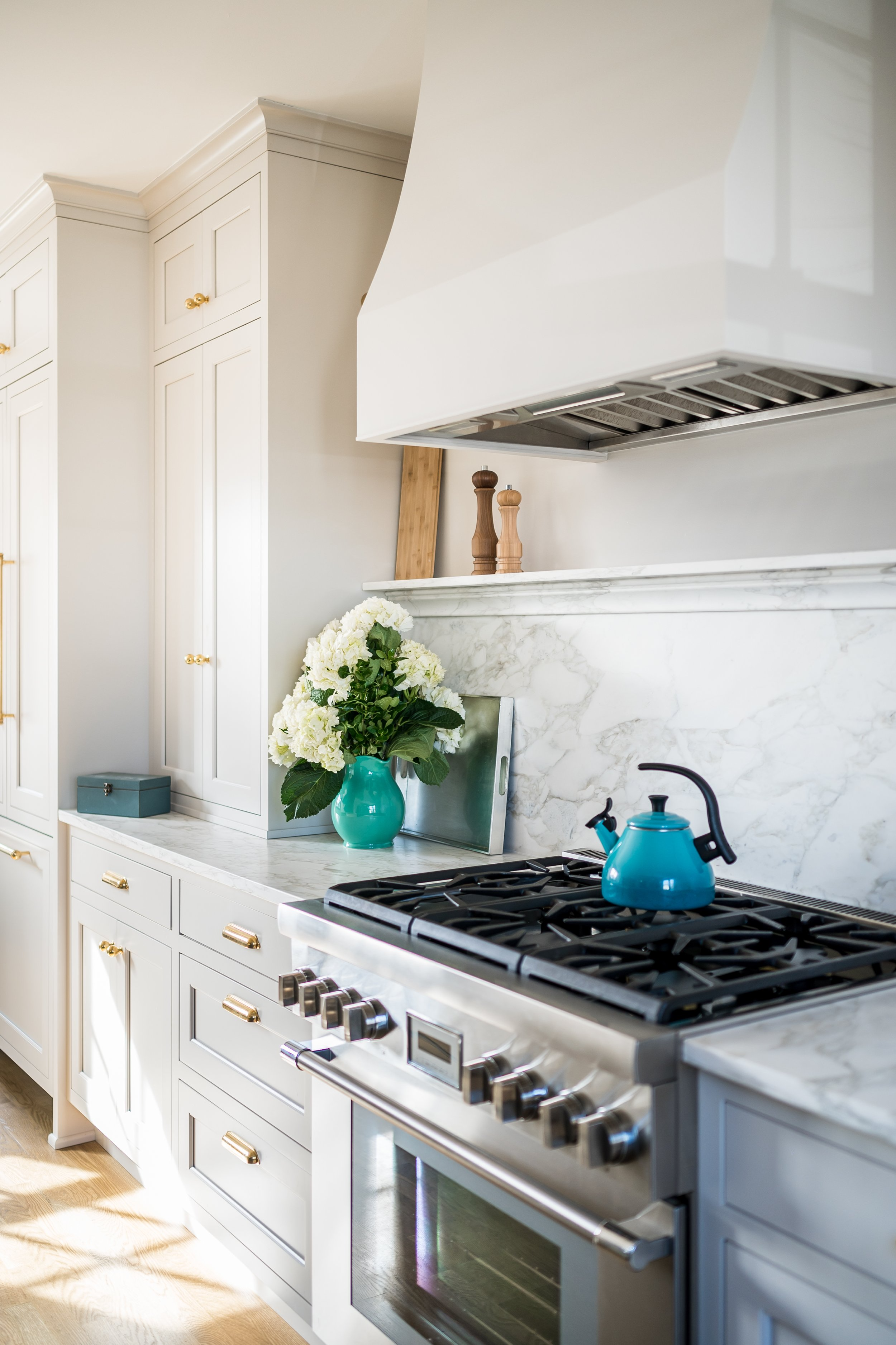 A modern kitchen with white cabinetry, a stainless steel stove, a vase with white hydrangeas, a tray, and a blue teapot on the countertop.