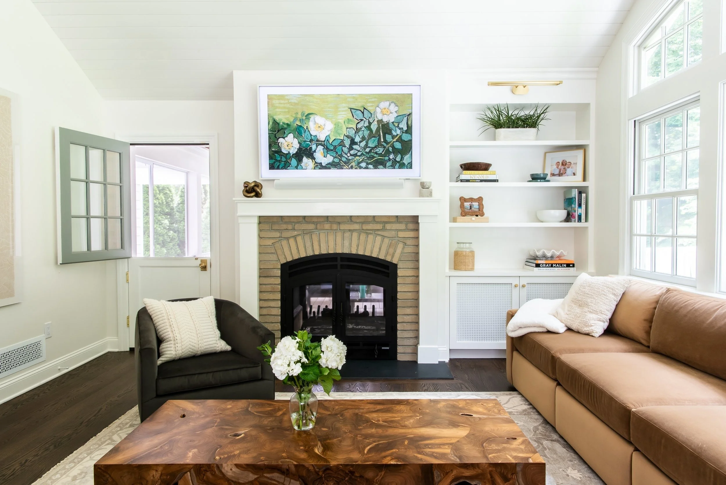 Living room with a brick fireplace, white built-in shelves, a framed painting above the fireplace, a brown sofa, a black armchair with a white pillow, a wooden coffee table with a vase of white flowers, large windows, and various decorative items.