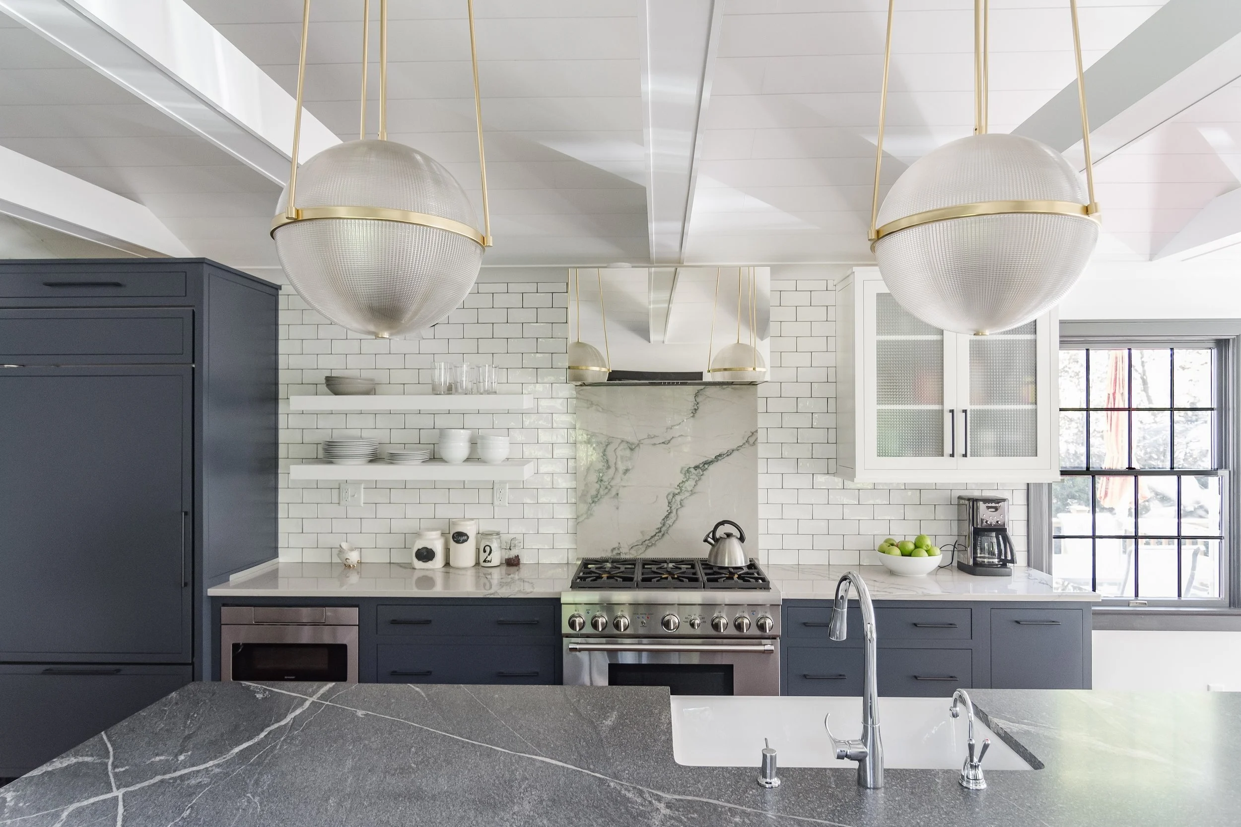 Modern kitchen with white subway tile backsplash, dark blue cabinets, marble accents, and two large ceiling light fixtures, with a window on the right and a kitchen island in the foreground.