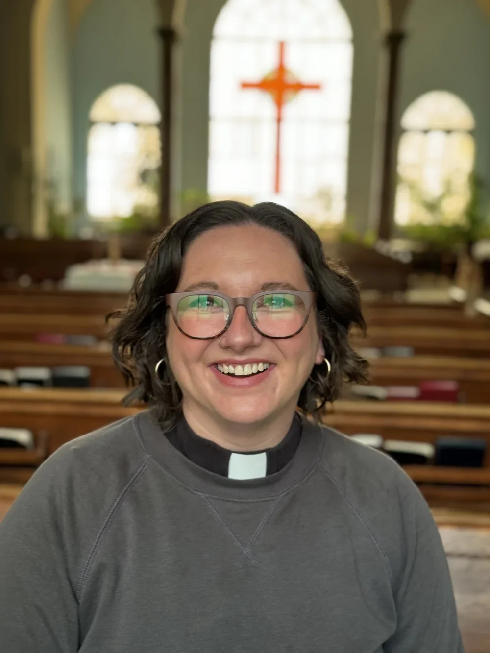 Lindsey is a white woman with glasses and short brown hair, wearing a clerical collar and gray sweatshirt