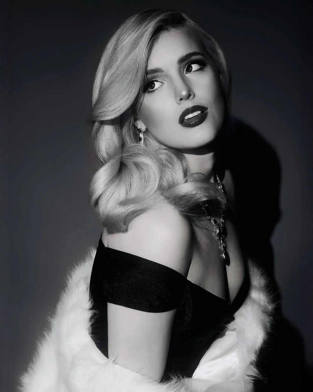 Black-and-white portrait of a woman with styled blonde hair, dark lipstick, and jewelry, wearing a fur stole and a black gown with an off-the-shoulder design.