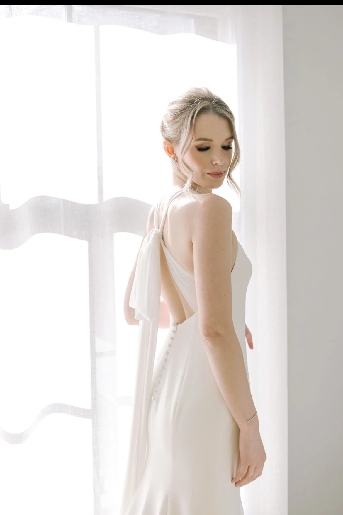 A bride in a white wedding gown standing near a window with sheer curtains, looking down with a gentle expression.