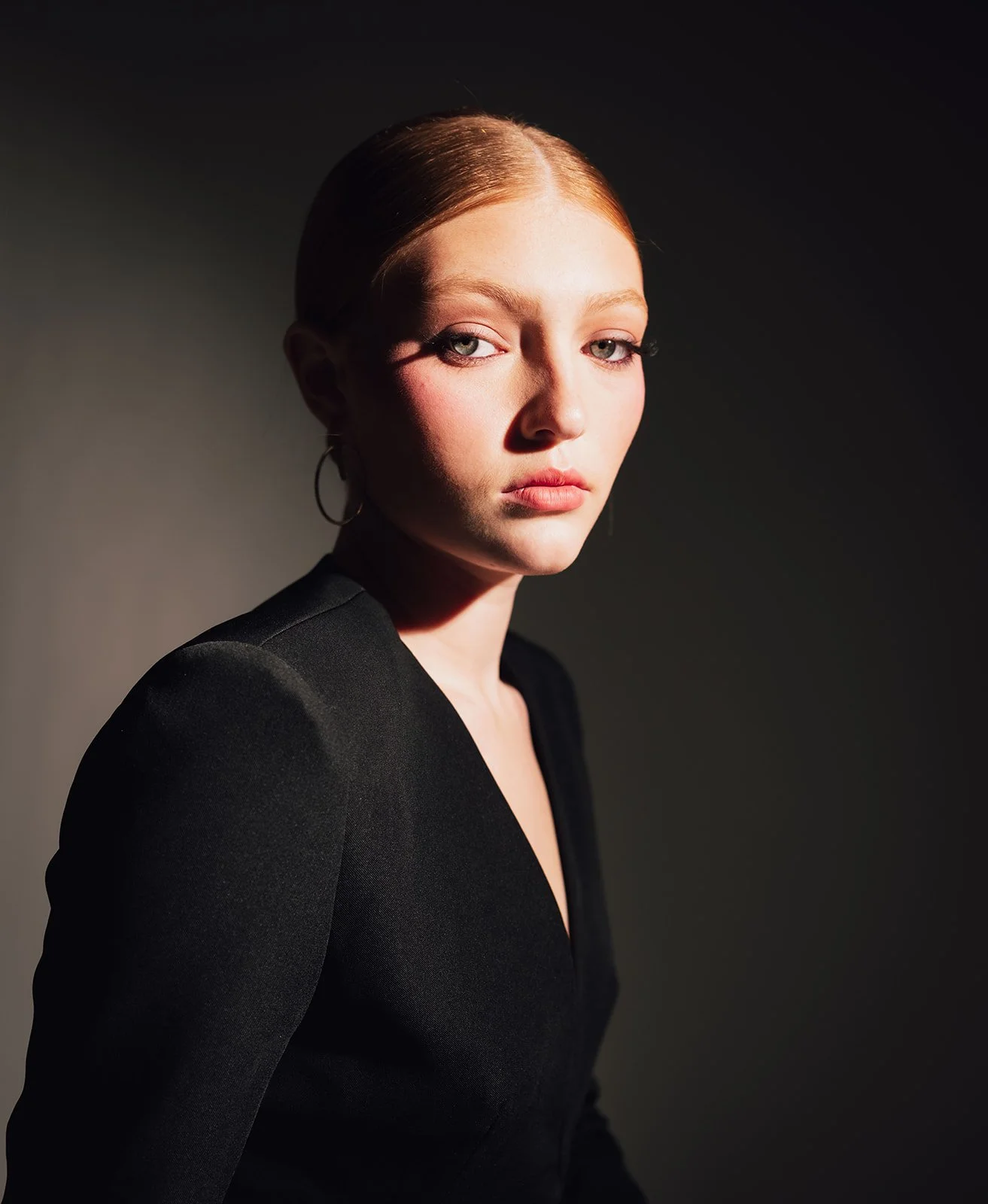 A portrait of a woman with red hair, wearing a black top, and looking into the camera with a serious expression, against a dark background.