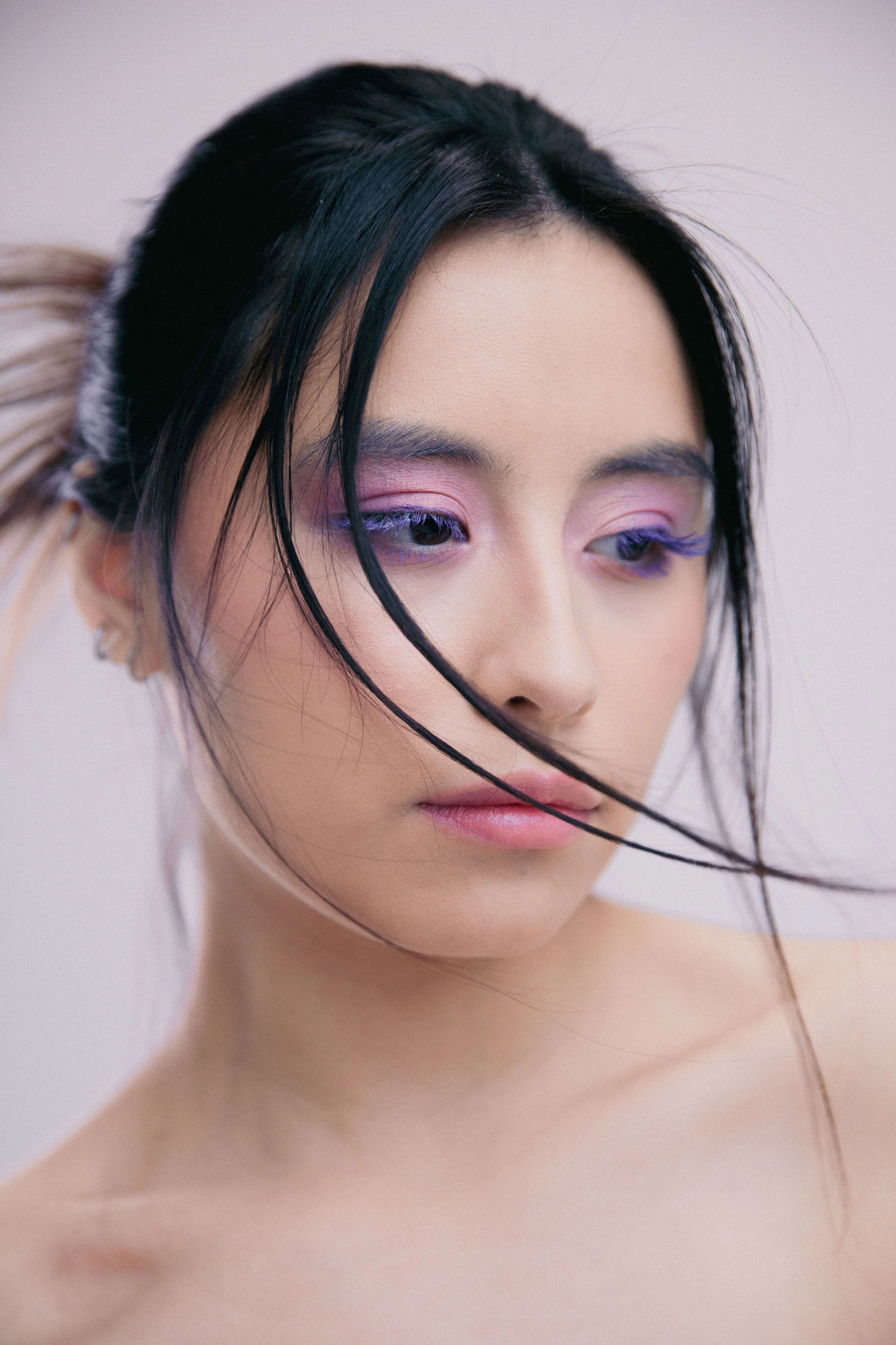 A close-up portrait of a young woman with long black hair, light skin, and pastel purple eyeshadow, looking downward with a gentle expression.