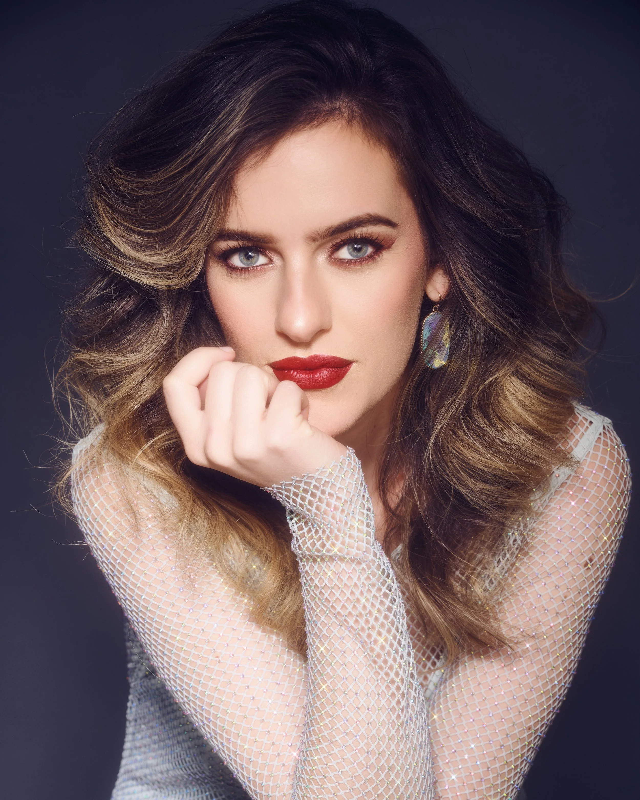 A woman with wavy brown hair and piercing blue eyes gazes directly at the camera. She has bold red lipstick and is wearing a sheer, glittery top and a colorful earring. Her pose suggests confidence, with her hand positioned near her face.