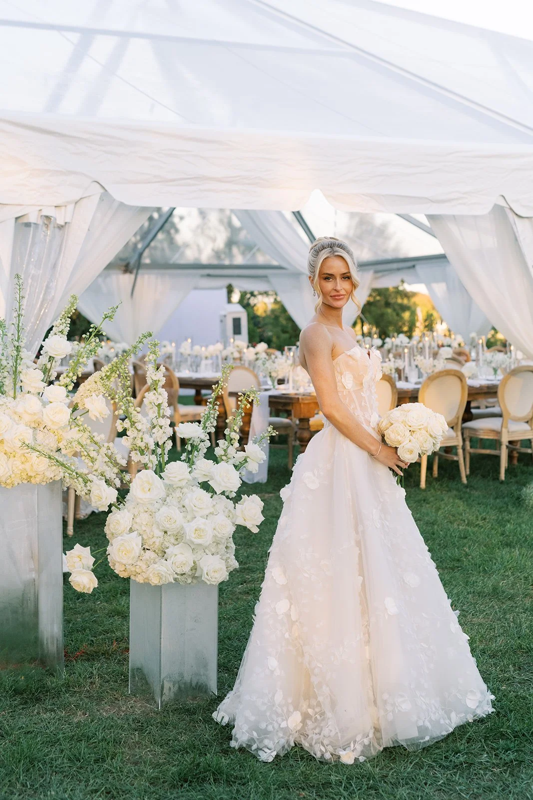 Bride standing in an outdoor tent surrounded by floral arrangements before guests arrive.
